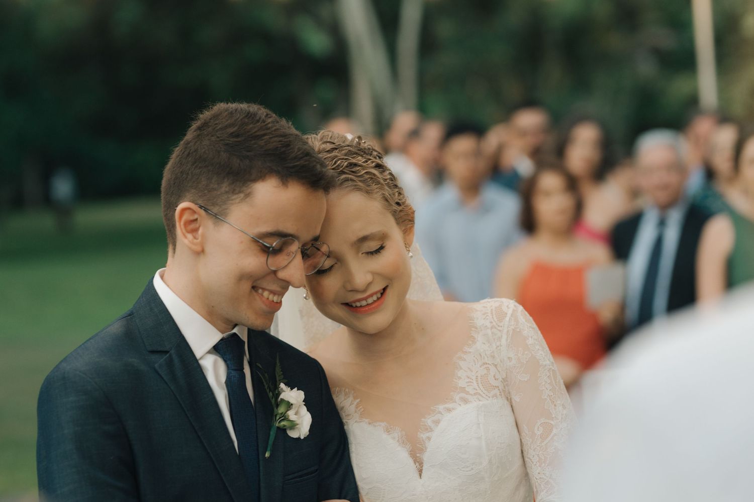 A tender moment between newlyweds as they share a laugh with wedding guests in the background.