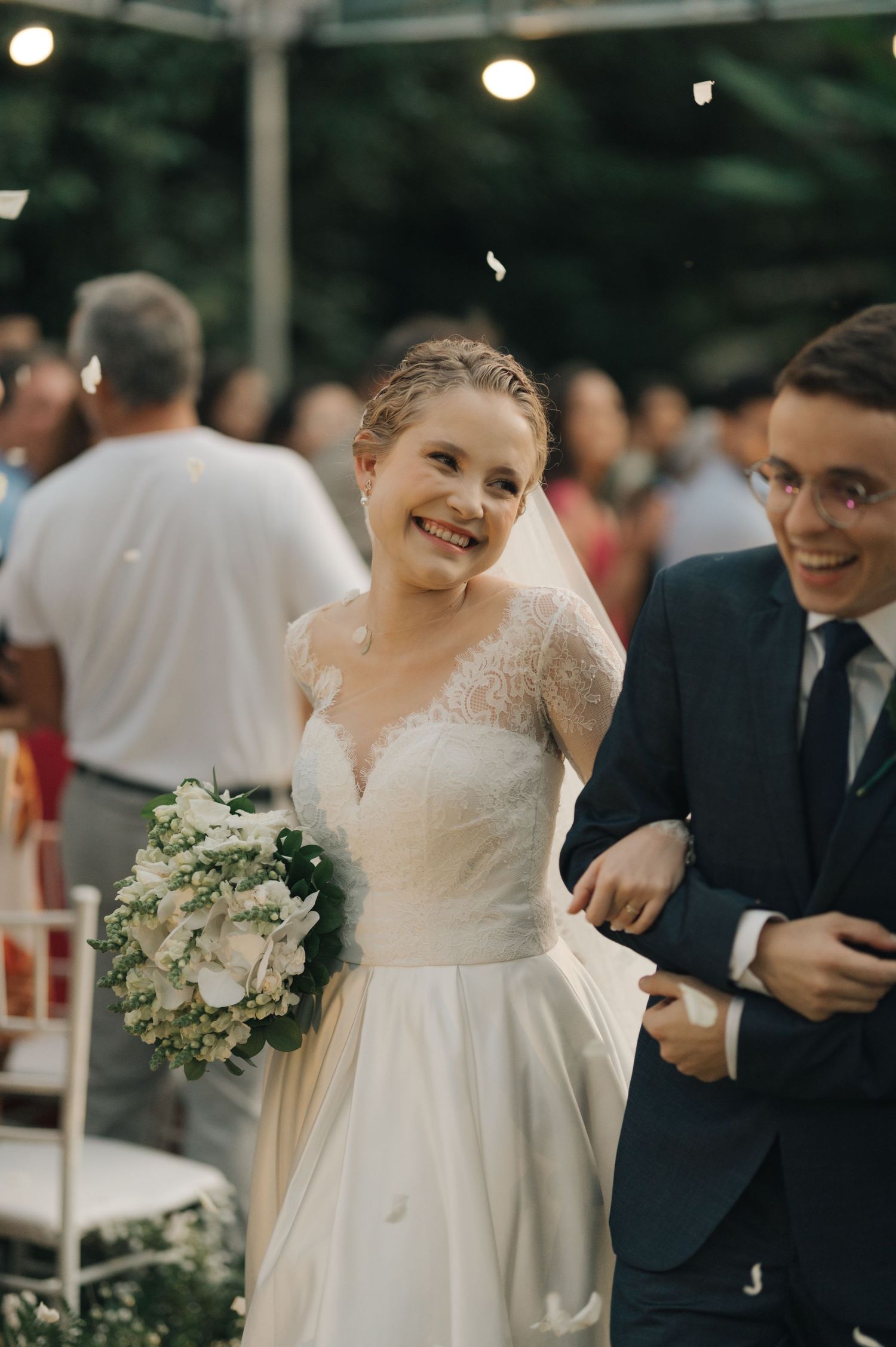 Bride holding white bouquet smiles radiantly beside groom at outdoor wedding reception.