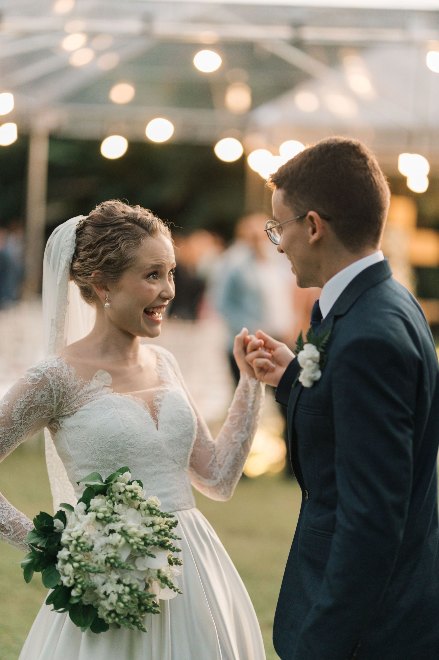 Bride with white bouquet and lace dress laughs with groom under outdoor reception lights.