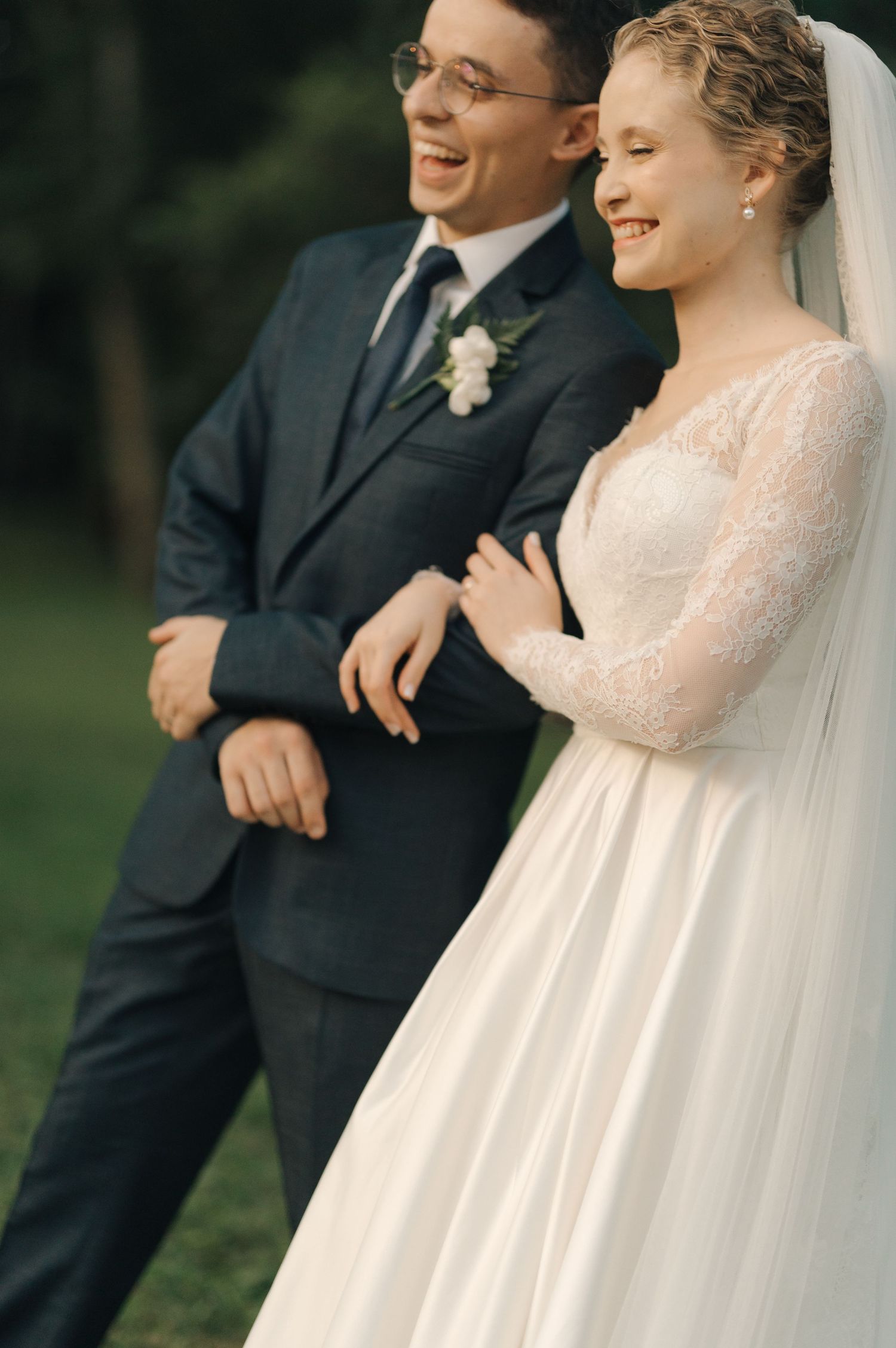 A joyful wedding couple shares a candid moment outdoors, with the bride in lace and groom in a dark suit.