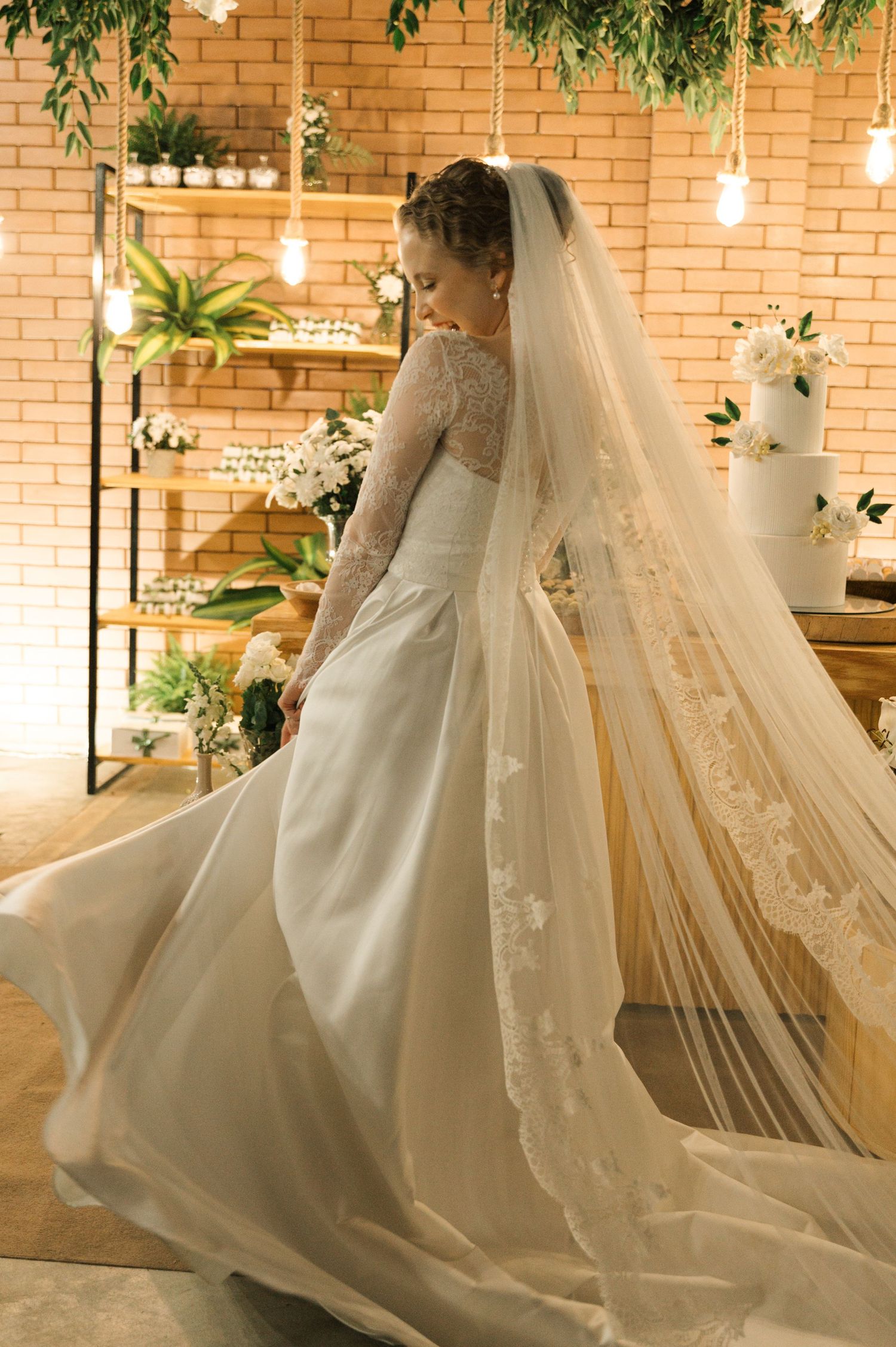 An elegant bride poses in a flowing lace wedding gown with a dramatic cathedral veil against a brick backdrop.