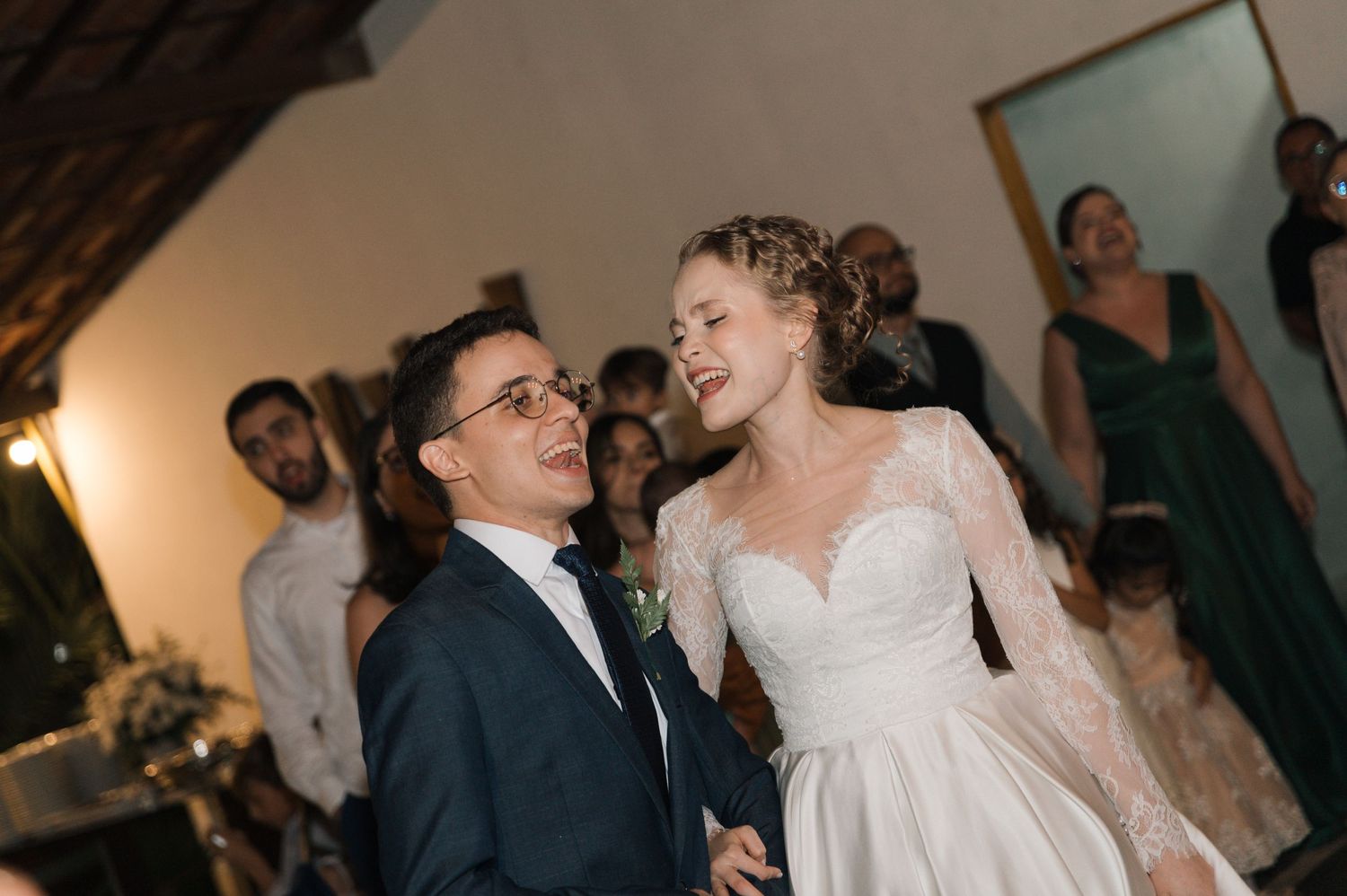 Wedding couple shares hearty laughter during their reception celebration with guests in background.