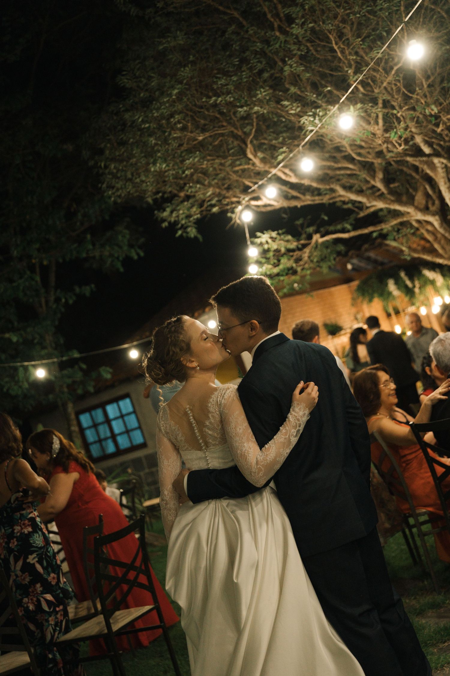 A romantic evening wedding kiss under string lights and trees in an outdoor garden setting.