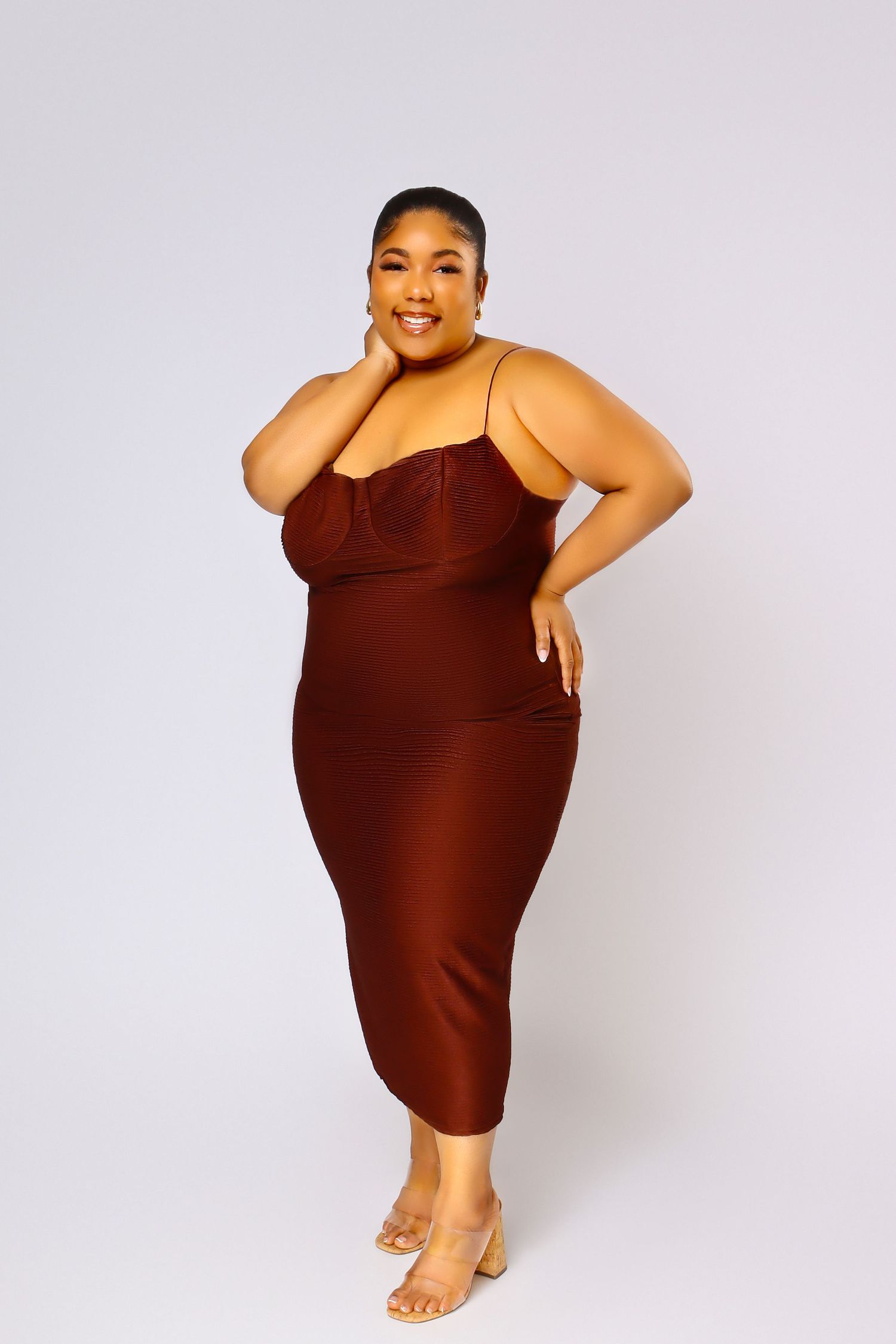Confident model in brown fitted dress poses with hand on hip against light background.