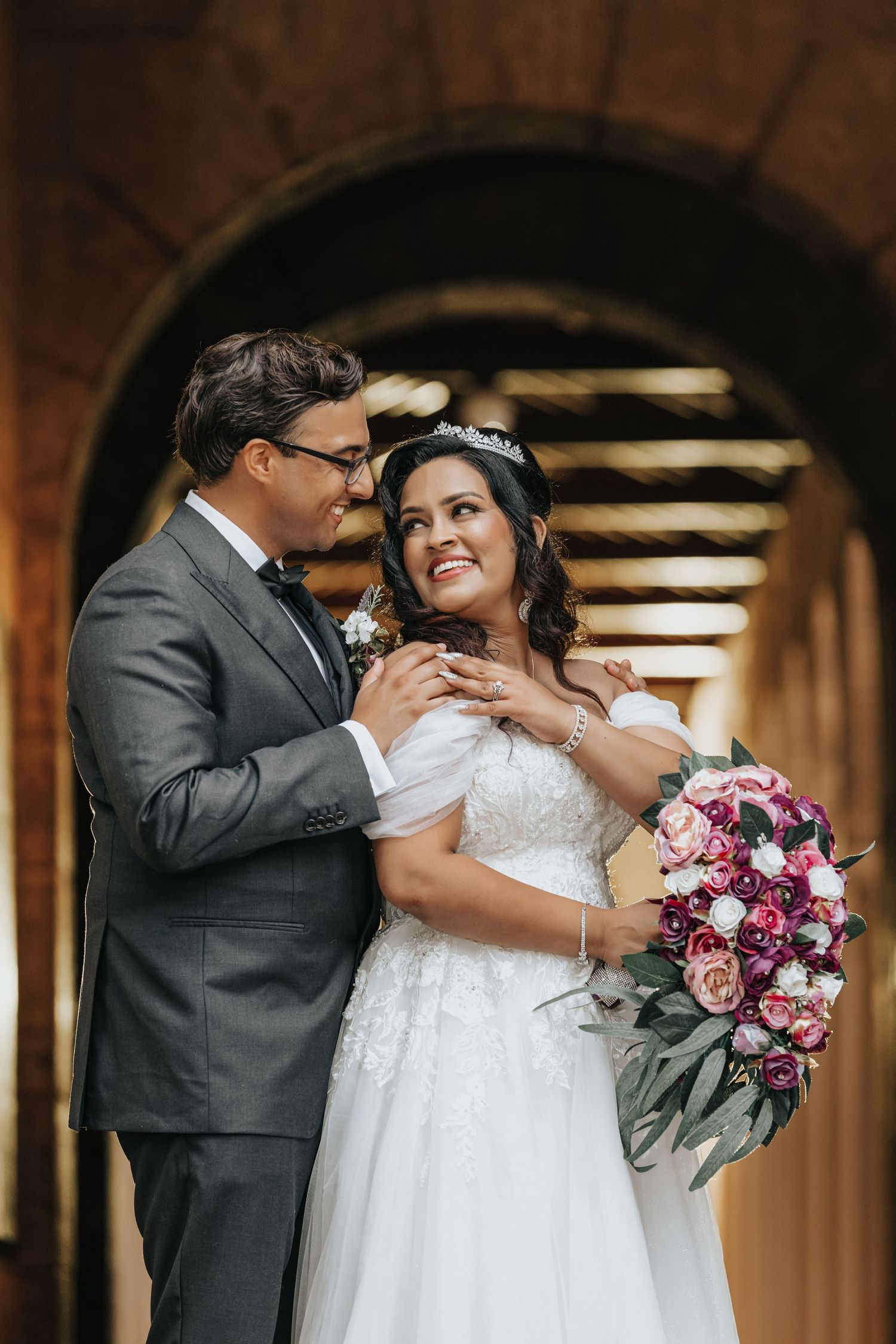A wedding couple poses under an archway with a pink and purple bouquet.