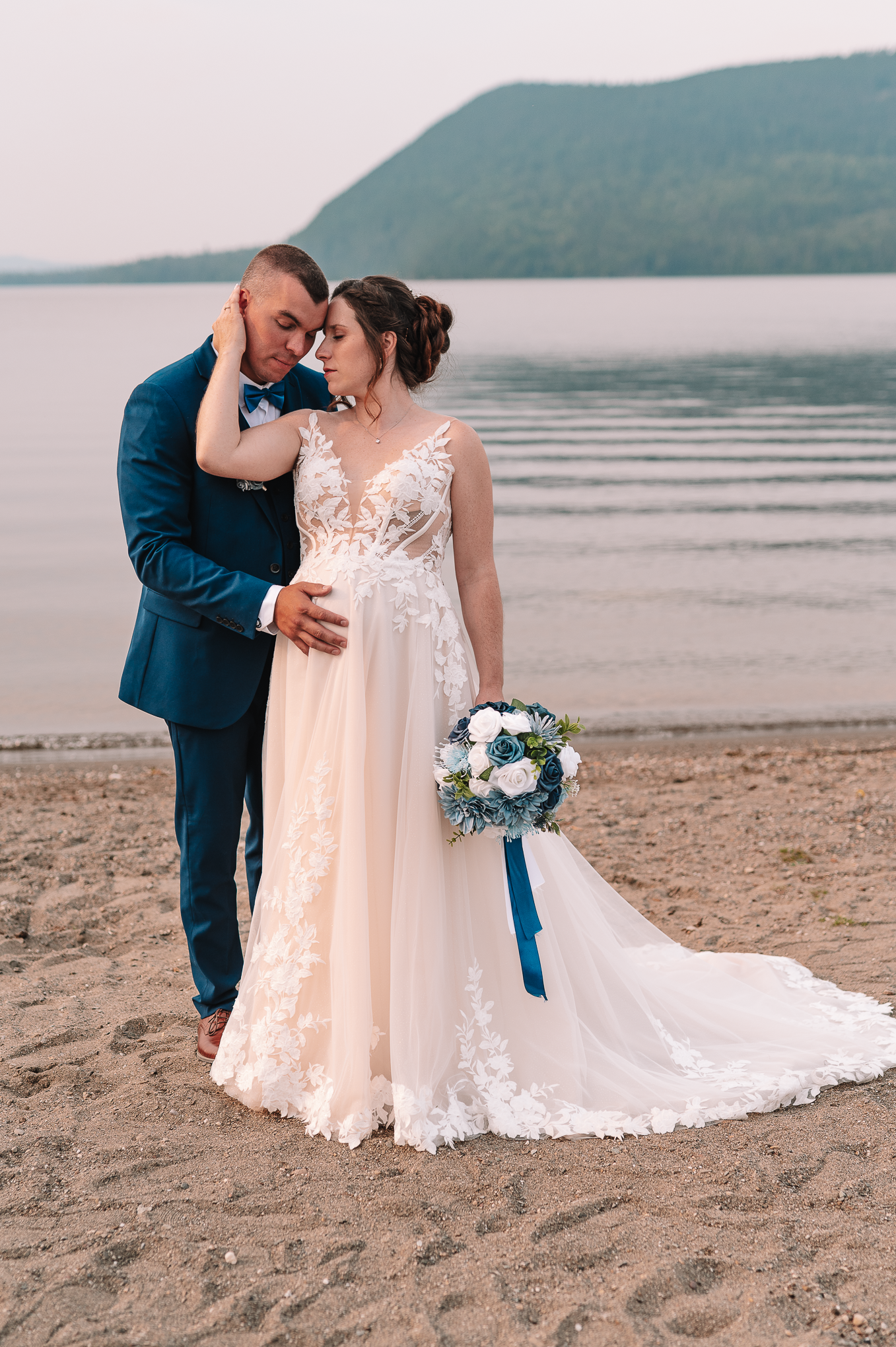 Romantic wedding couple embracing on shoreline in formal attire.
