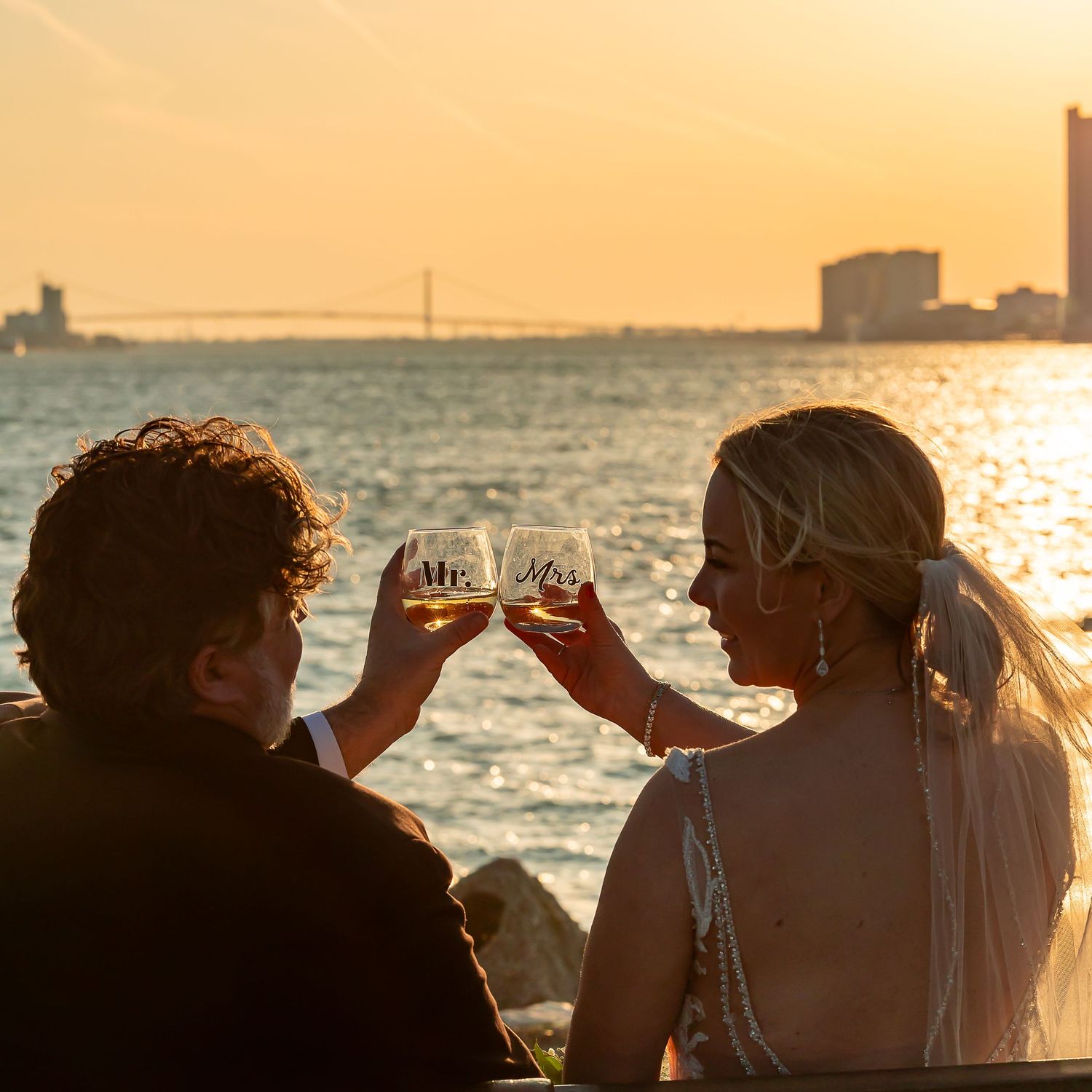 A couple toasts with wine glasses against a golden sunset over the water.