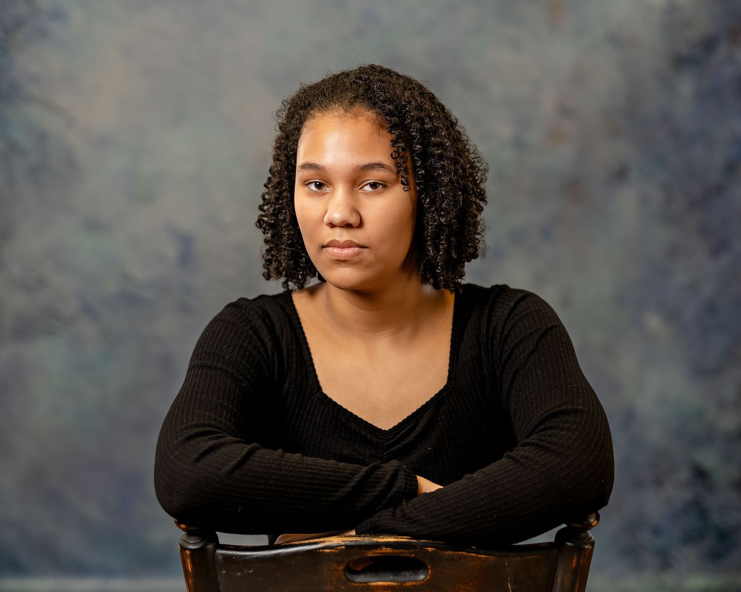 Young woman with natural curly hair sits with arms crossed on a chair against a mottled blue-gray studio backdrop.