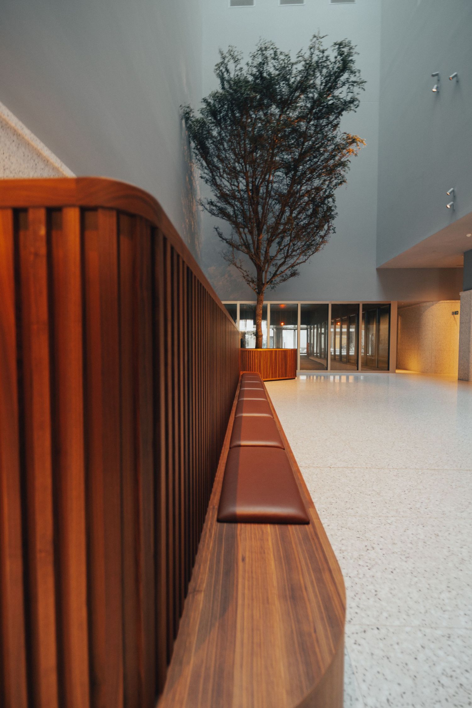 Moderne hal met houten balustrade en hoge binnenboom tegen grijze muur.