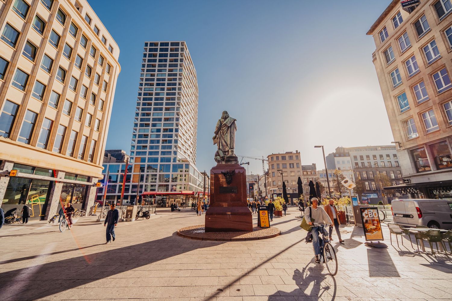 Stadsplein met hoge moderne gebouwen en historisch standbeeld in het centrum.