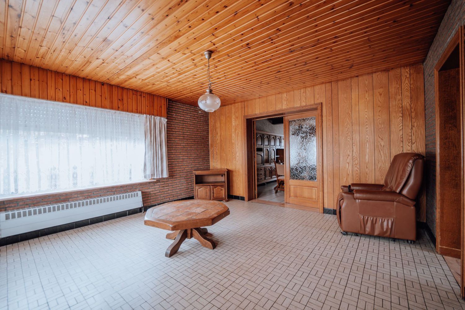 Retro woonkamer met houten lambrisering, bruine leren stoel en grote ramen.