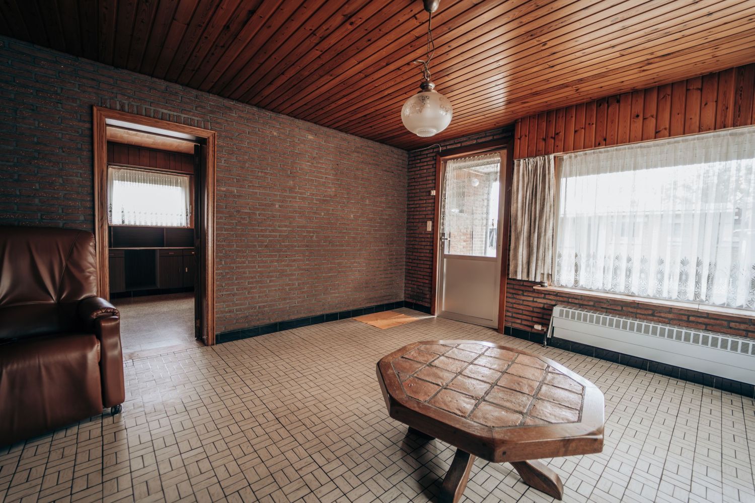 Ruime kamer met houten plafond, ronde tafel en grote ramen die natuurlijk licht binnenlaten.