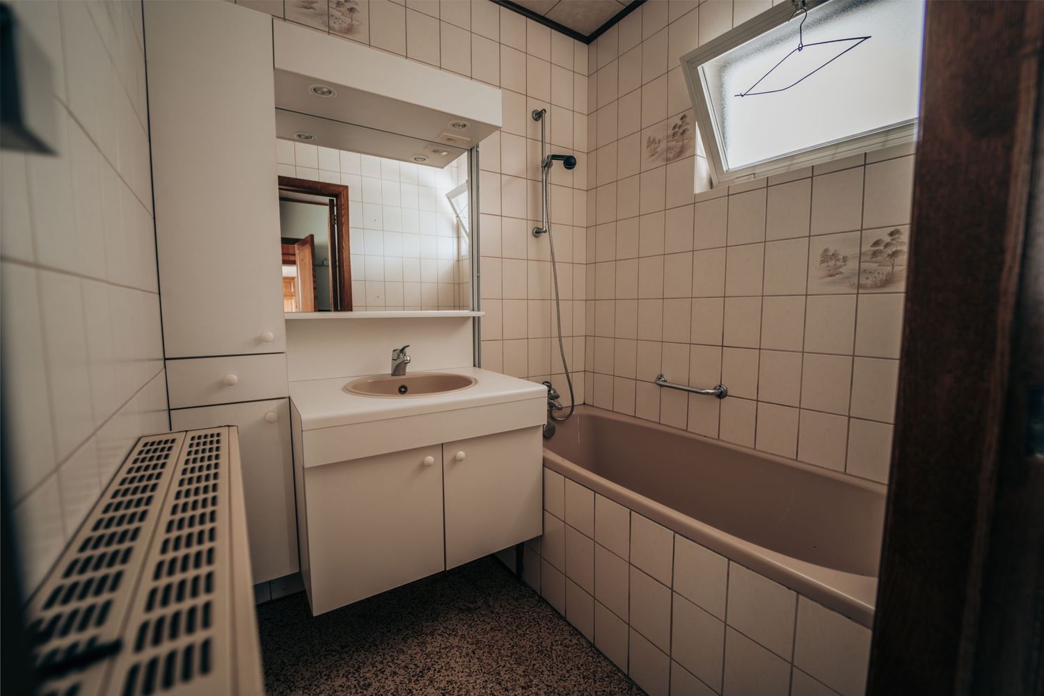 Een gedateerd badkamerinterieur met witte tegelwanden, een ingebouwd bad en een vintage wastafelmeubel.