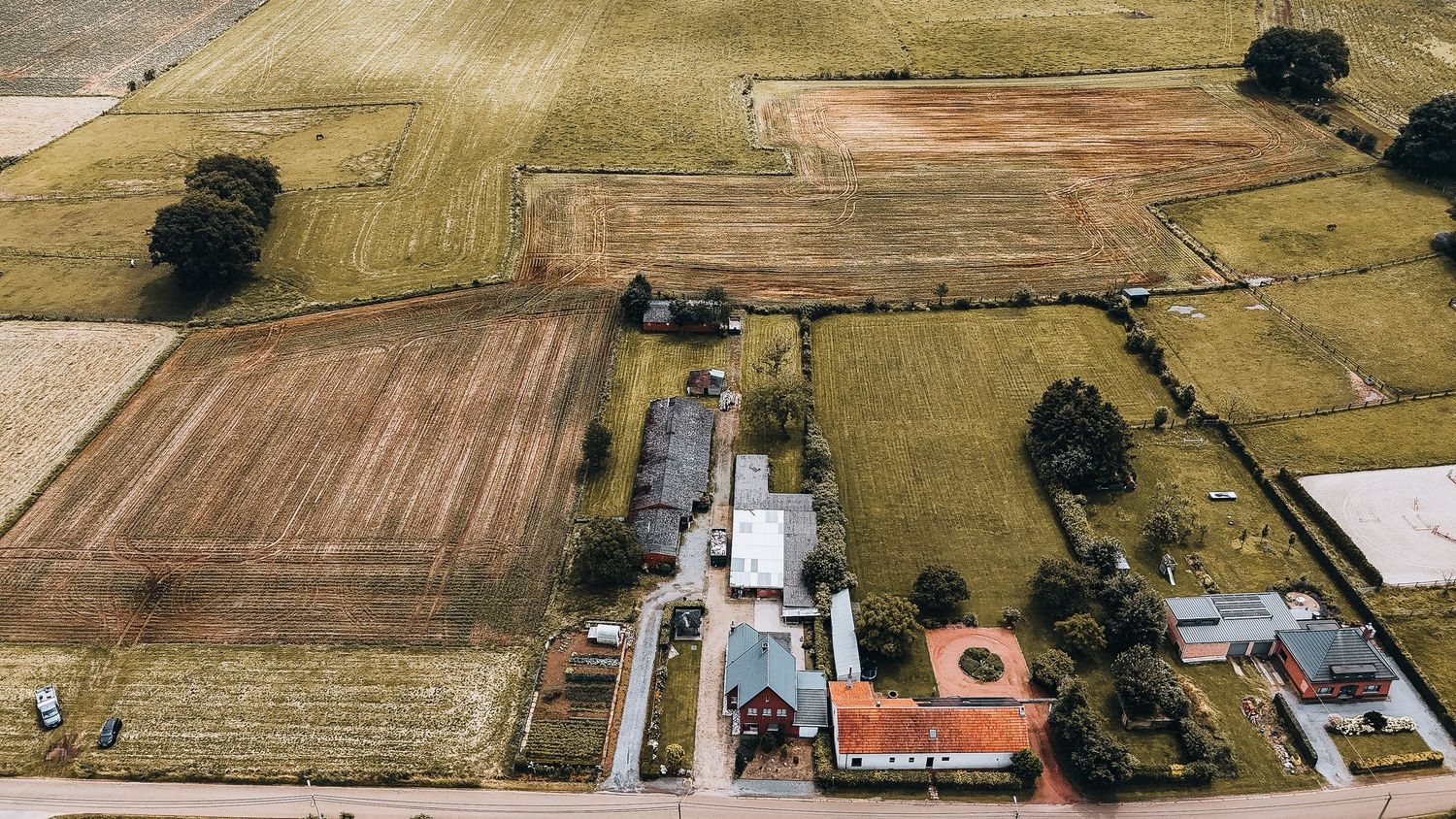 Drone-opname van akkerland met gebouwen en een cirkelvormig sportveld tussen gecultiveerde velden.