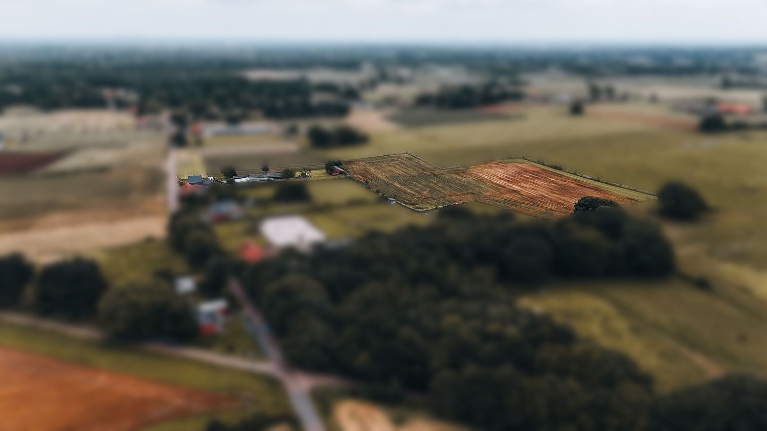 Tilt-shift luchtfoto van landelijk boerenland met gebouwen en velden die een dromerig effect creëren.