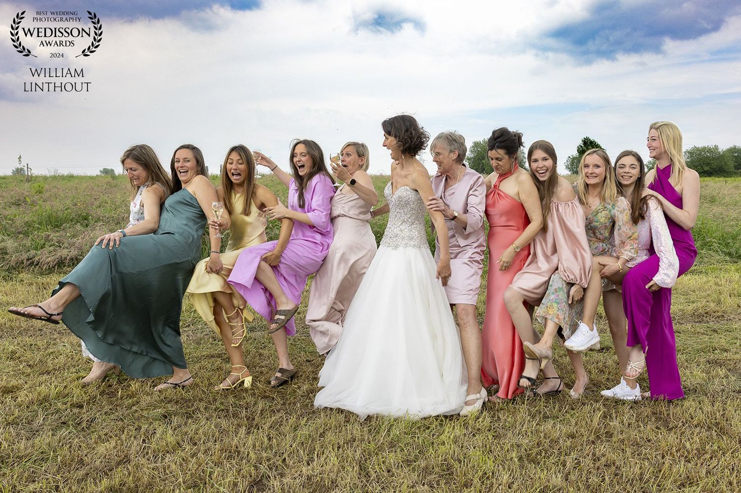 A bridal party in colorful dresses poses playfully on a grassy hill under blue skies.