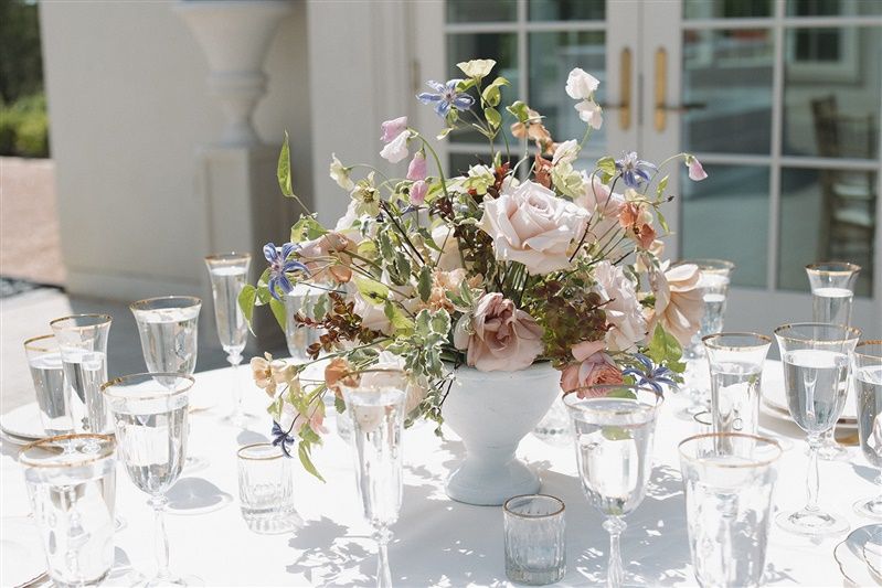 Elegant floral centerpiece with roses and greenery in a white vase on an outdoor table set with crystal glassware.