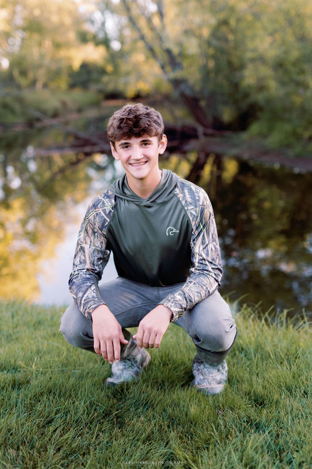 Person in camouflage sleeves and gray hoodie crouches on grass near a pond at sunset.