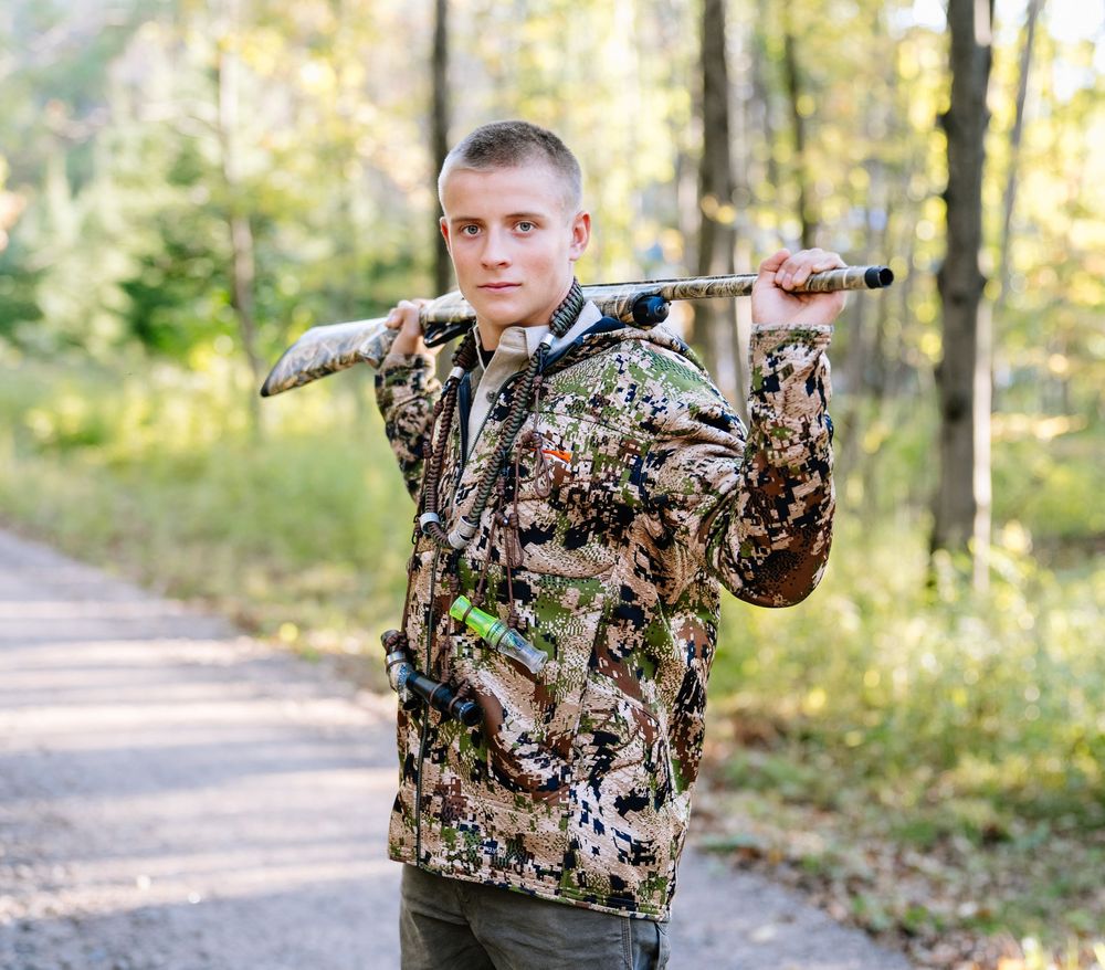 Person in camouflage hunting gear holding rifle across shoulders on forest path.
