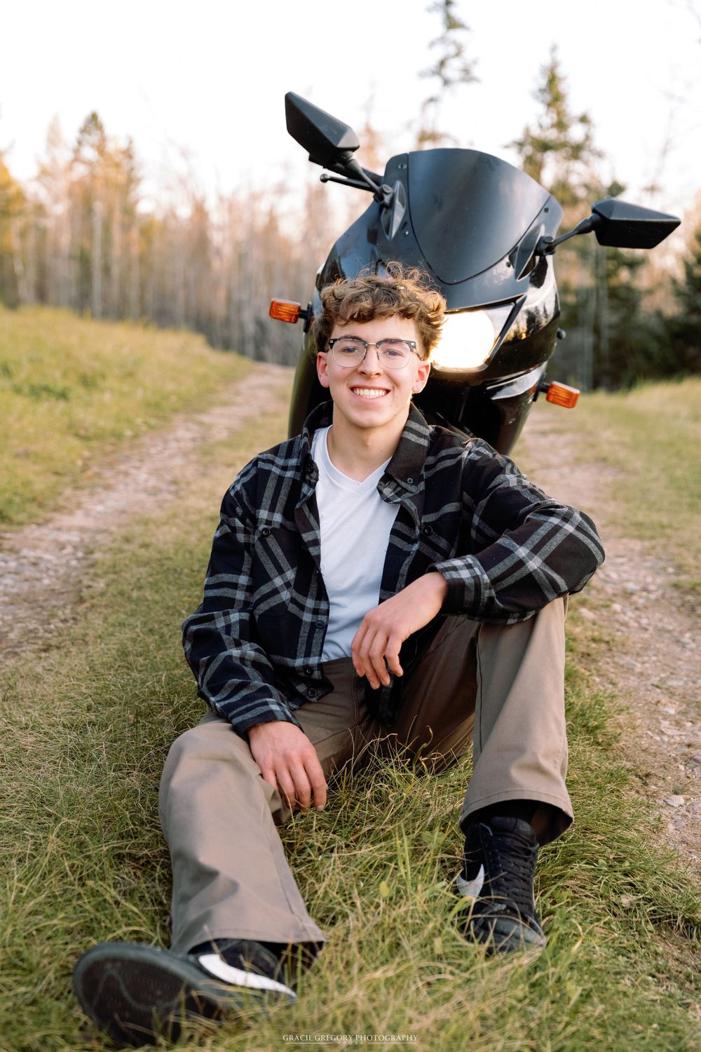 Person in plaid shirt poses with black motorcycle on dirt path through field.