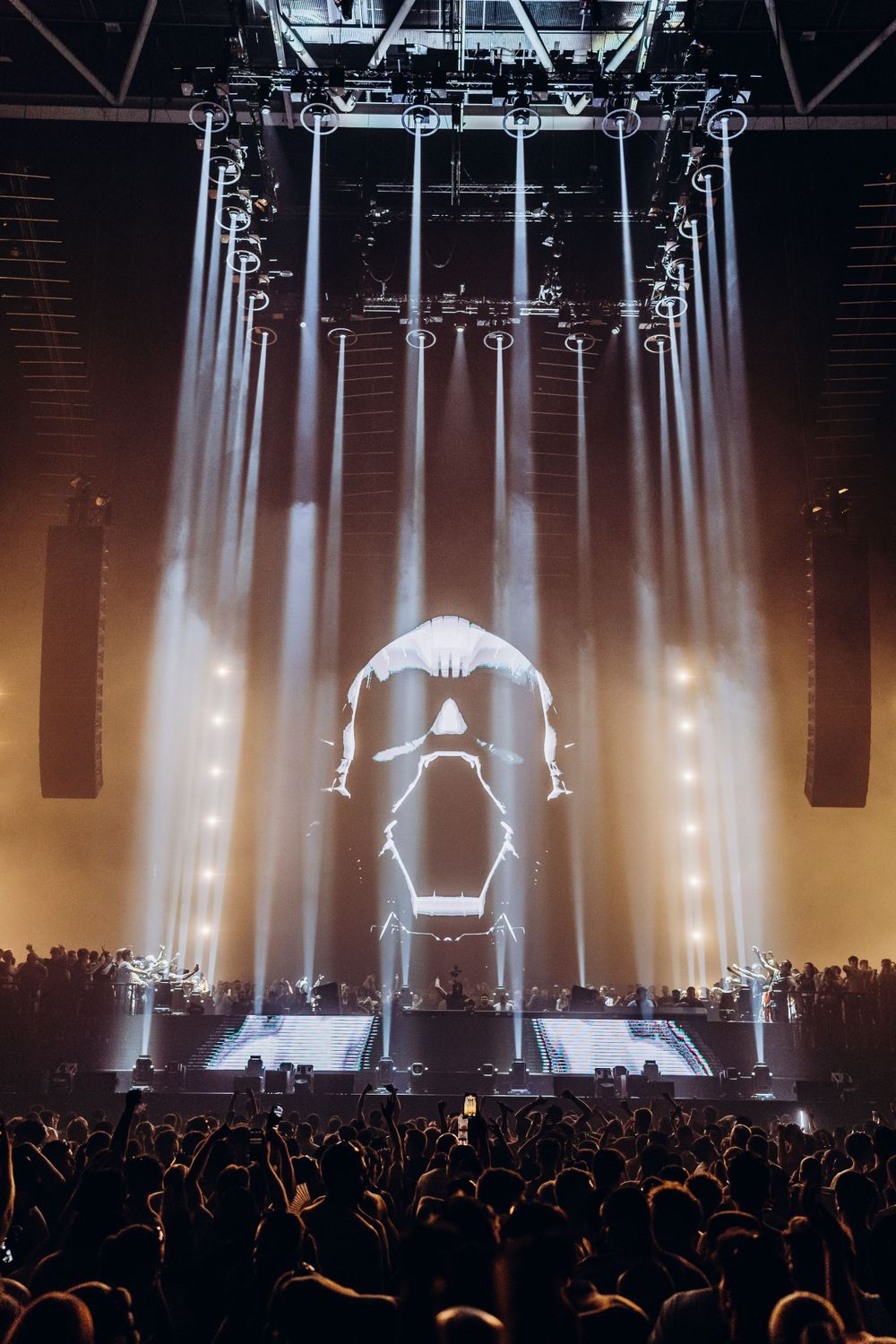 Music festival stage with dramatic white spotlights illuminating crowd in golden ambient lighting.