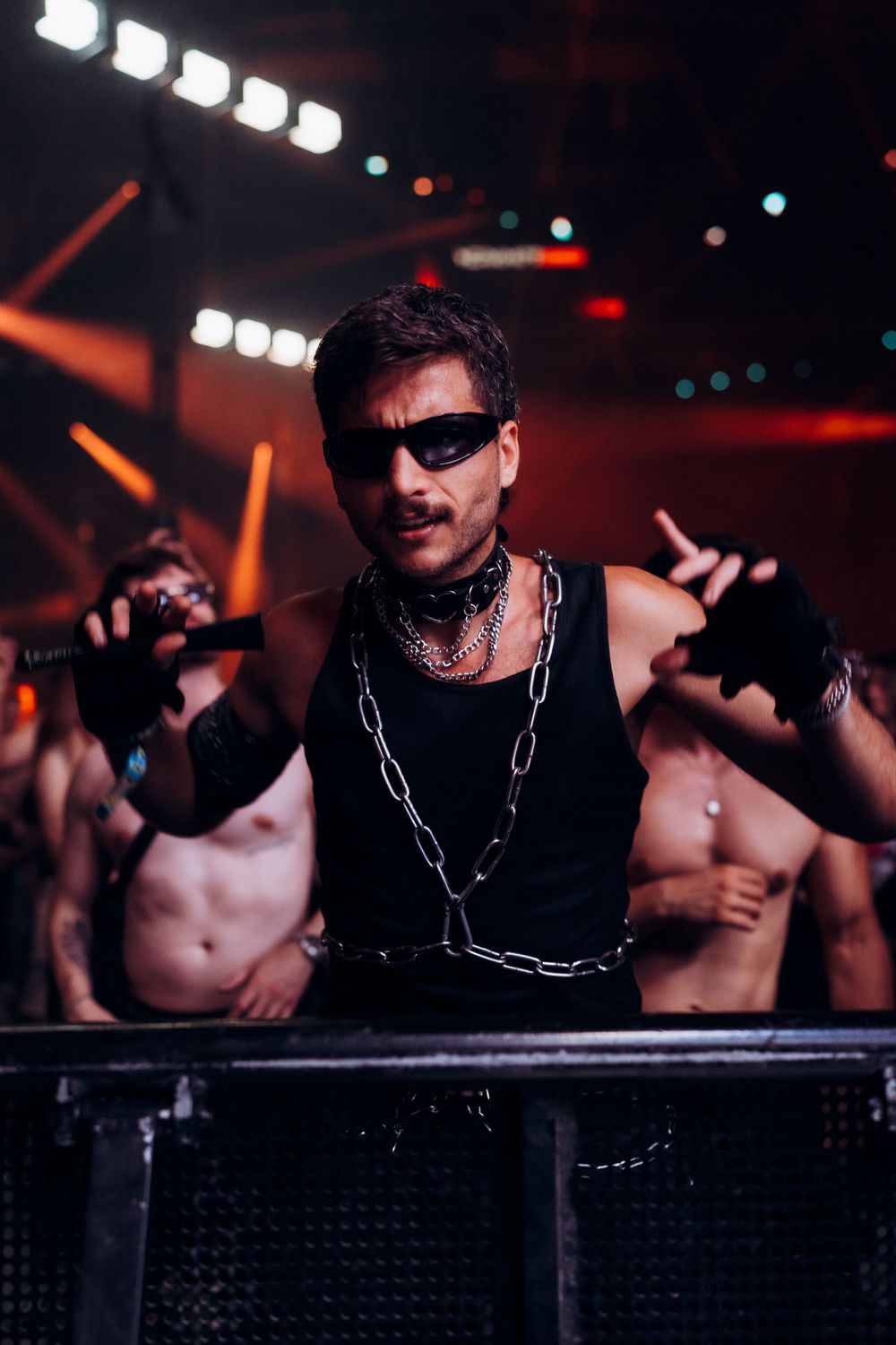 DJ performing at a nightclub wearing sunglasses and chains with dramatic red lighting and crowd in background.
