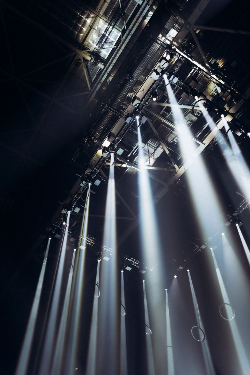 Dramatic white spotlights beaming down from concert venue ceiling with fog effects.