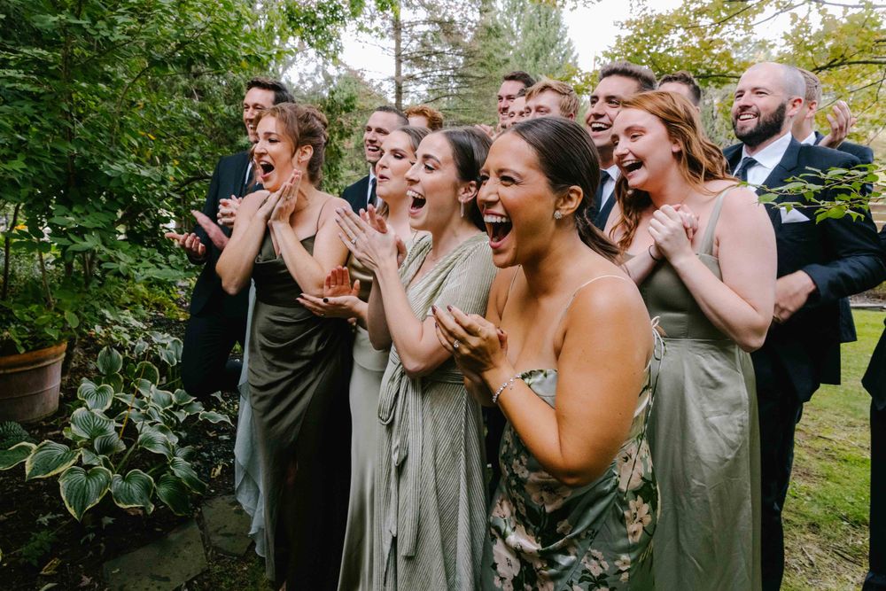 Bridesmaids in sage green dresses react with excitement during wedding celebration.