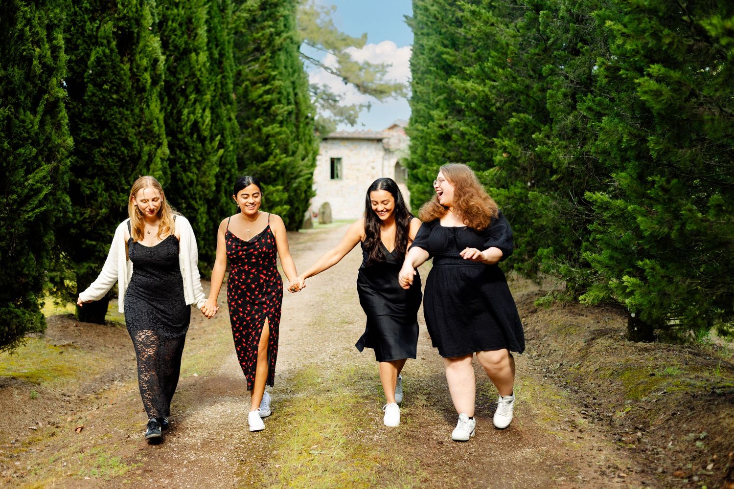 Friends walk together holding hands on a path between tall cypress trees.