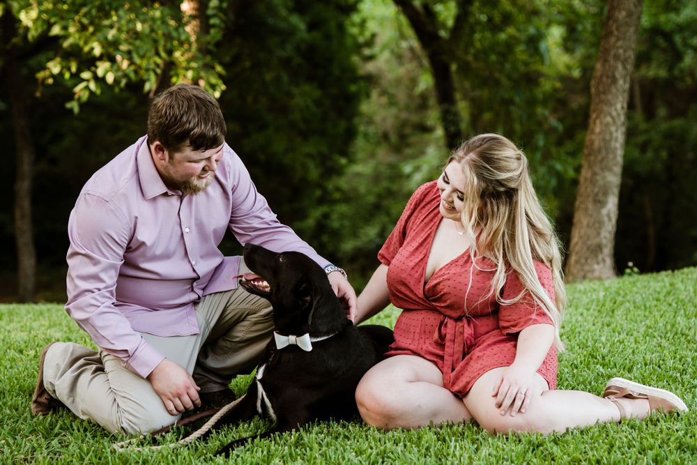 A couple sits on grass with their black dog enjoying a sunny day outdoors together.