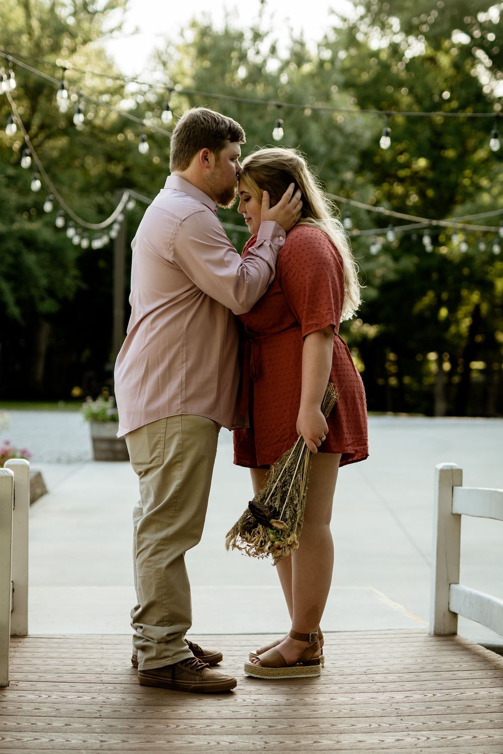 A romantic couple embraces on a wooden deck at sunset with string lights in background.