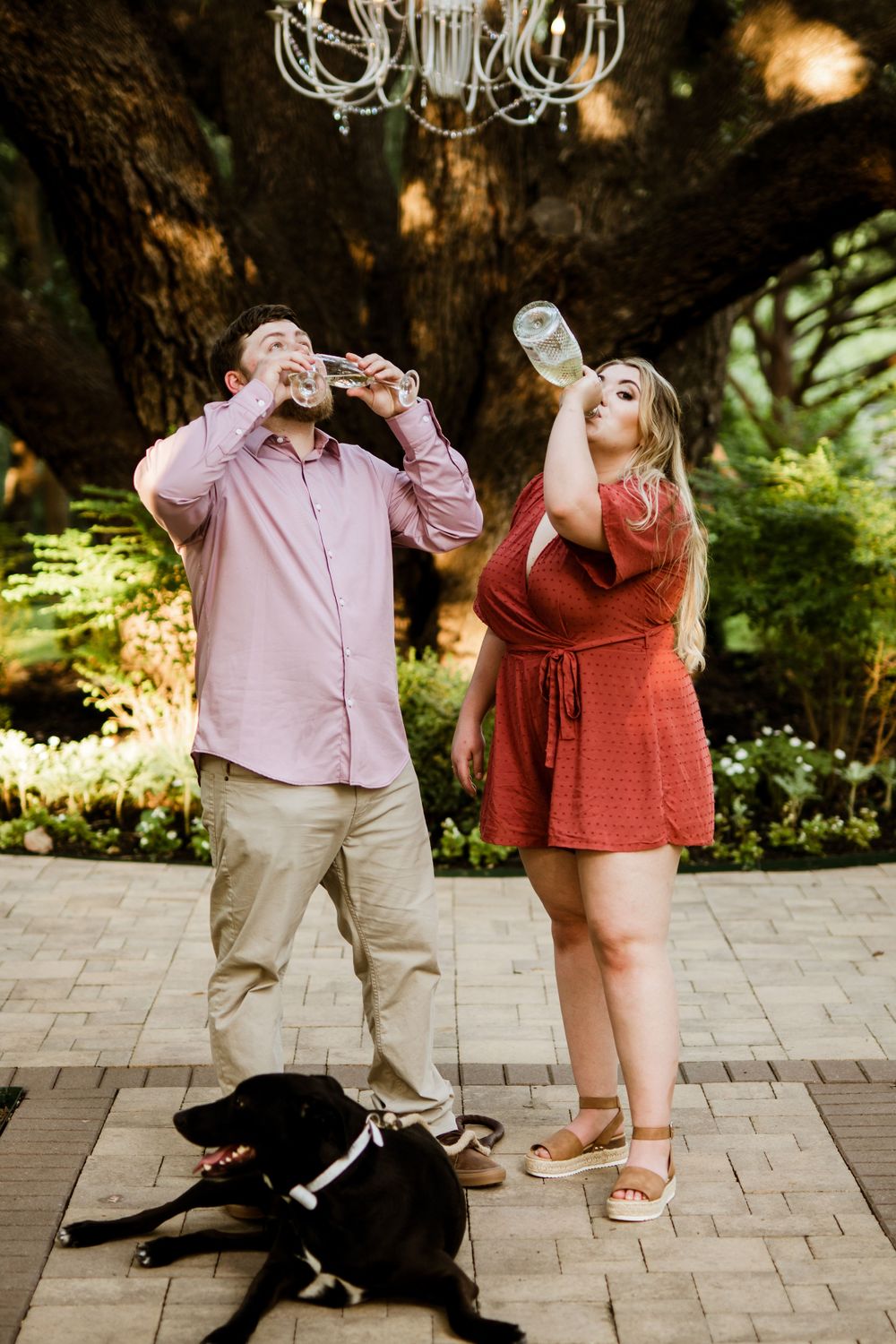 A playful couple drinks beverages with their black dog resting at their feet.