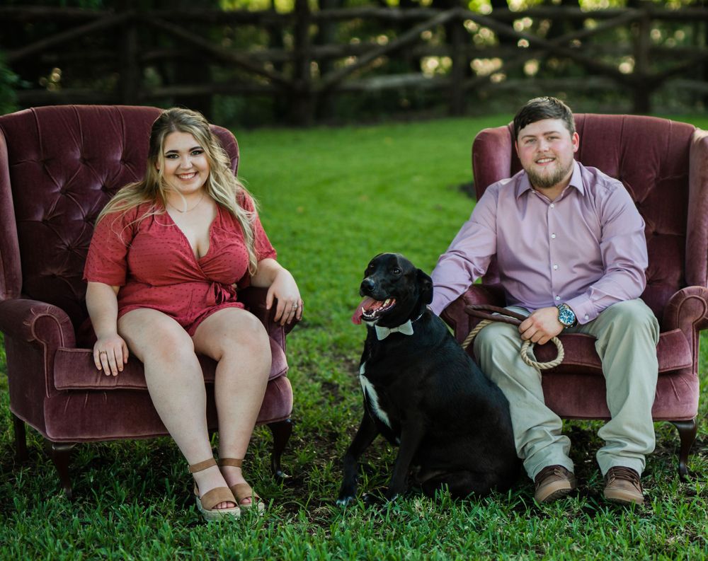 A couple sits with their black dog on velvet chairs in a garden setting.