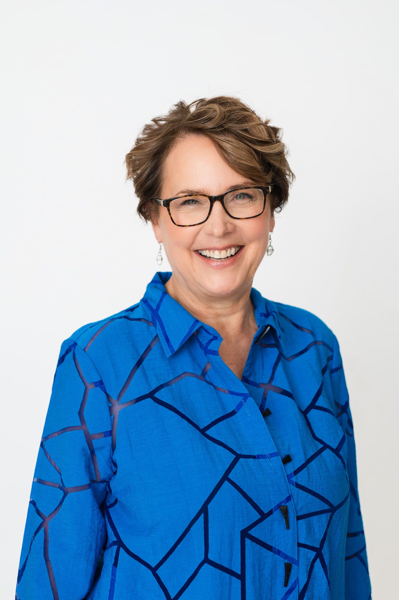 Professional wearing blue patterned blouse and glasses smiles warmly in studio portrait.