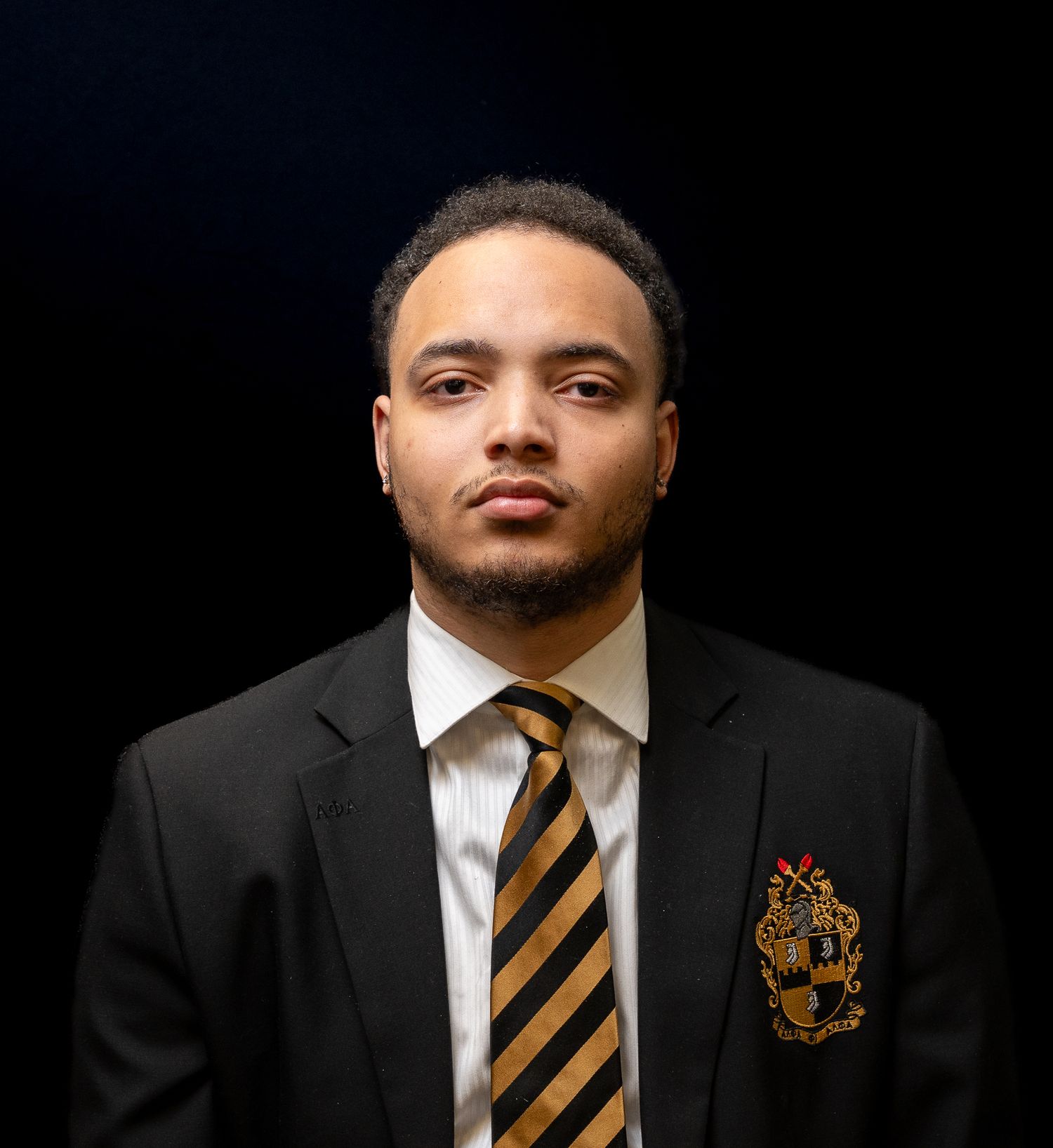 Professional portrait of someone wearing a dark suit with a striped tie and crest against a black background.