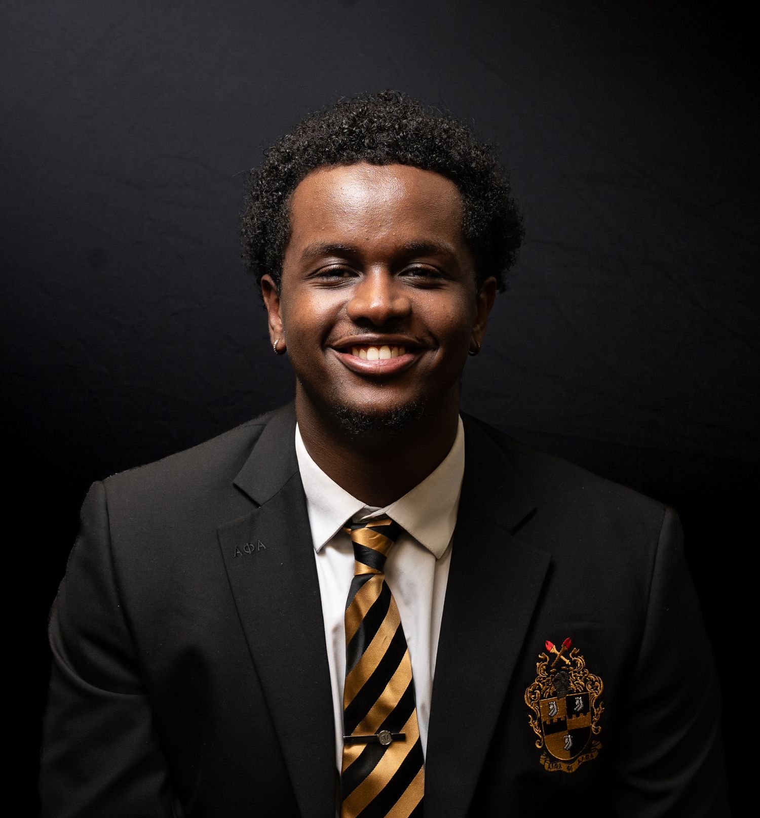 Professional portrait of someone in a dark suit with a striped tie and embroidered crest against a black background.
