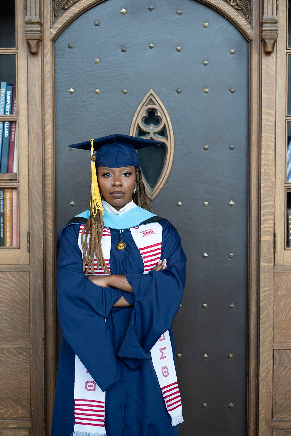 Graduate in blue cap and gown posing against ornate door with yellow tassel.