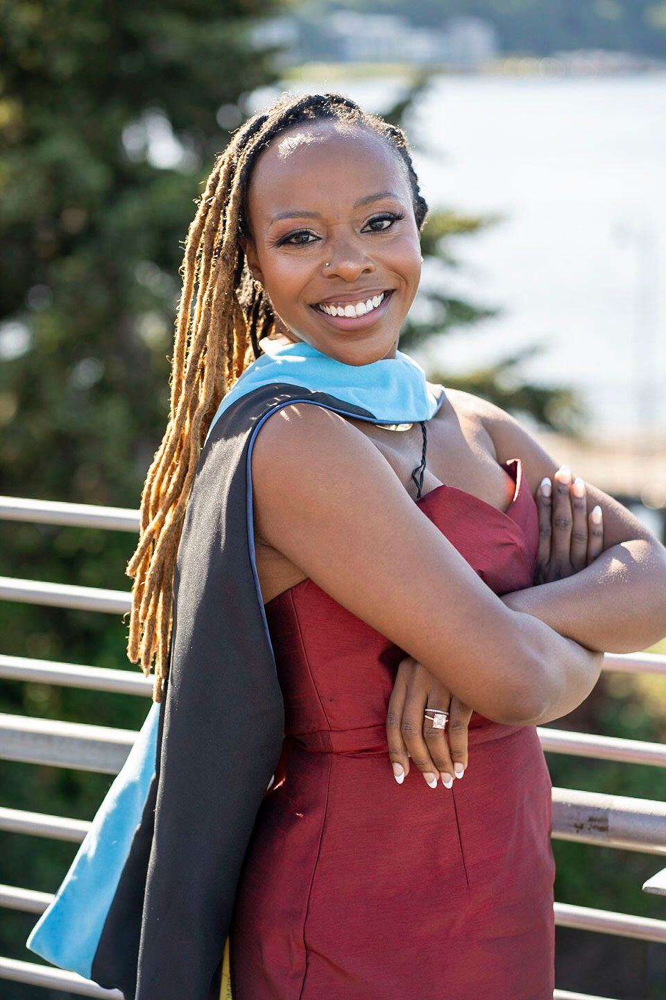 Graduate in burgundy dress and blue stole smiling outdoors on sunny day.