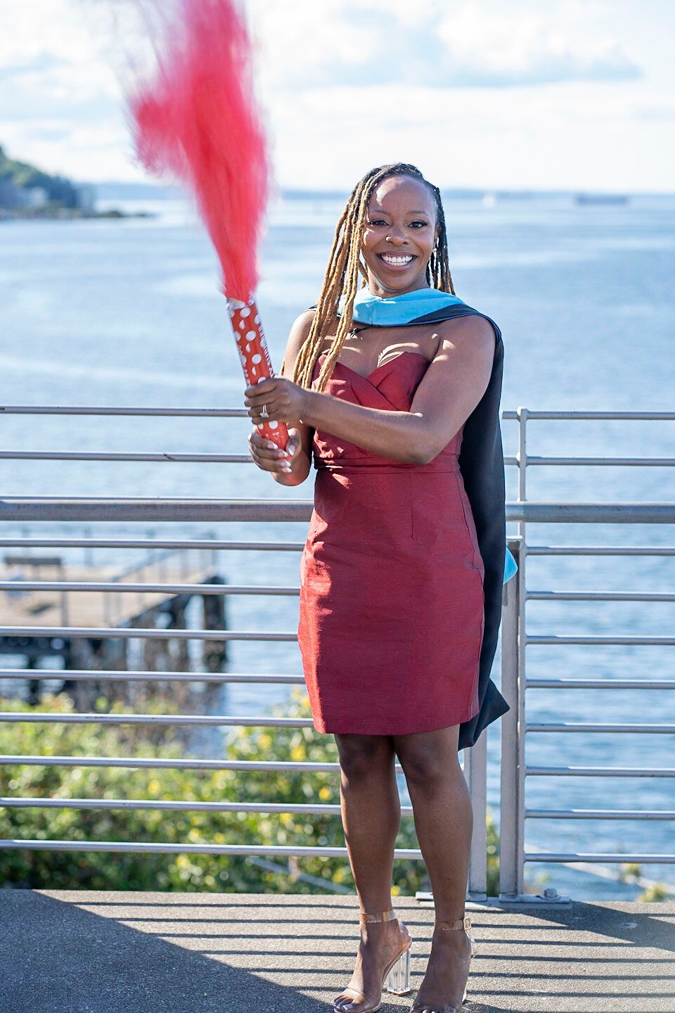 Graduate celebrates on waterfront deck holding red smoke flare while wearing burgundy dress and graduation stole.