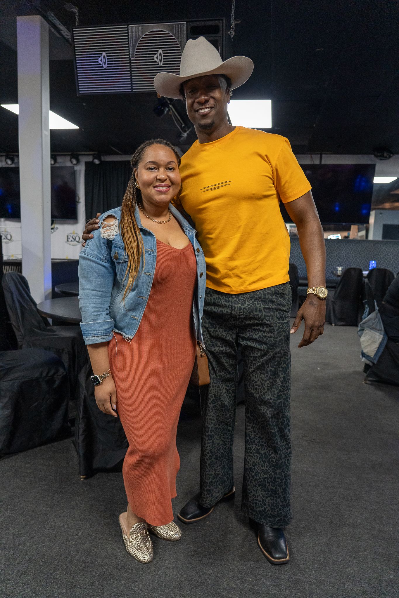 A couple poses together in an indoor venue, with the person in orange dress and another in a yellow shirt and cowboy hat.