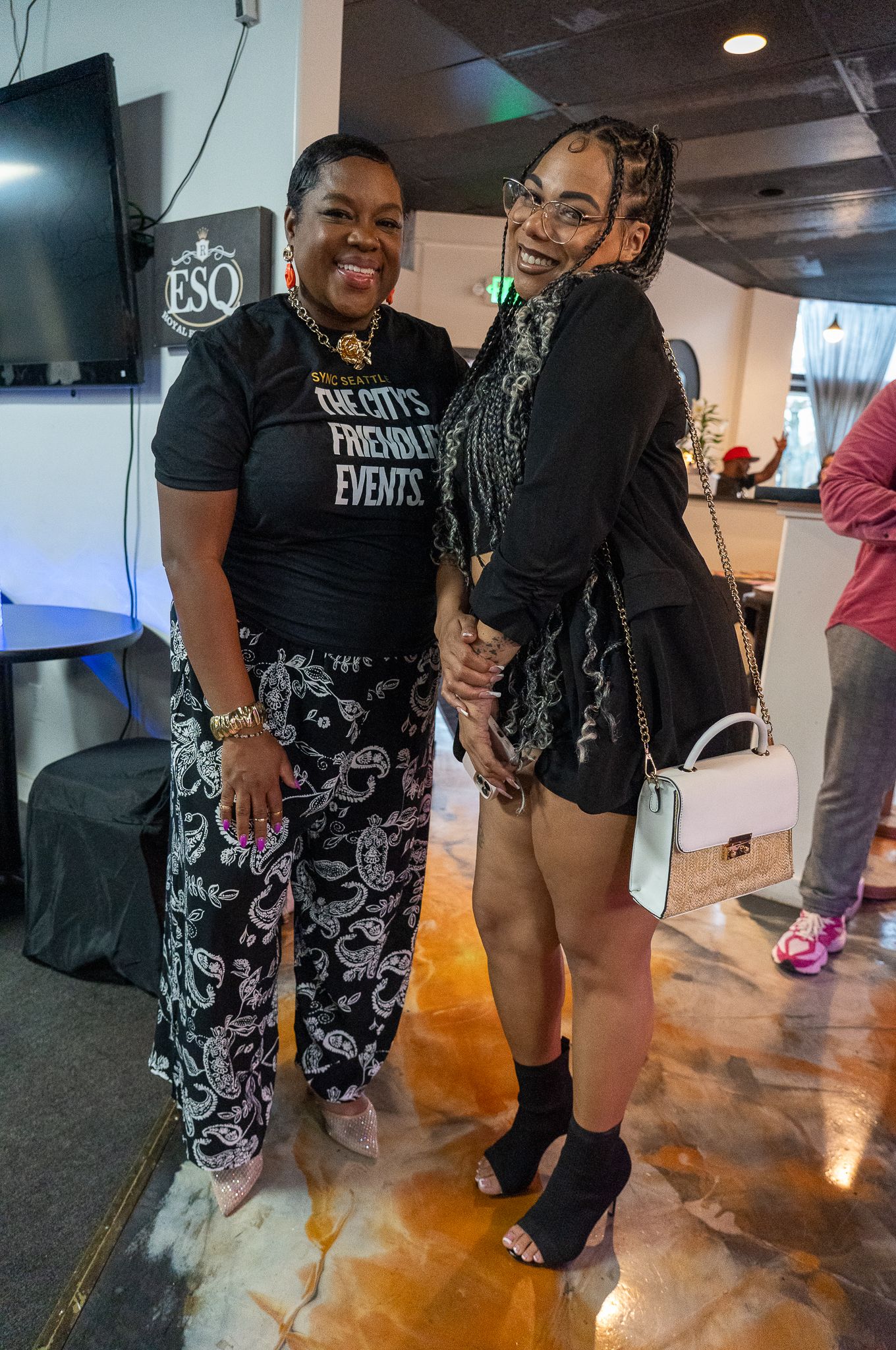 Two people posing together joyfully at an indoor event, with a TV mounted on the wall behind them.