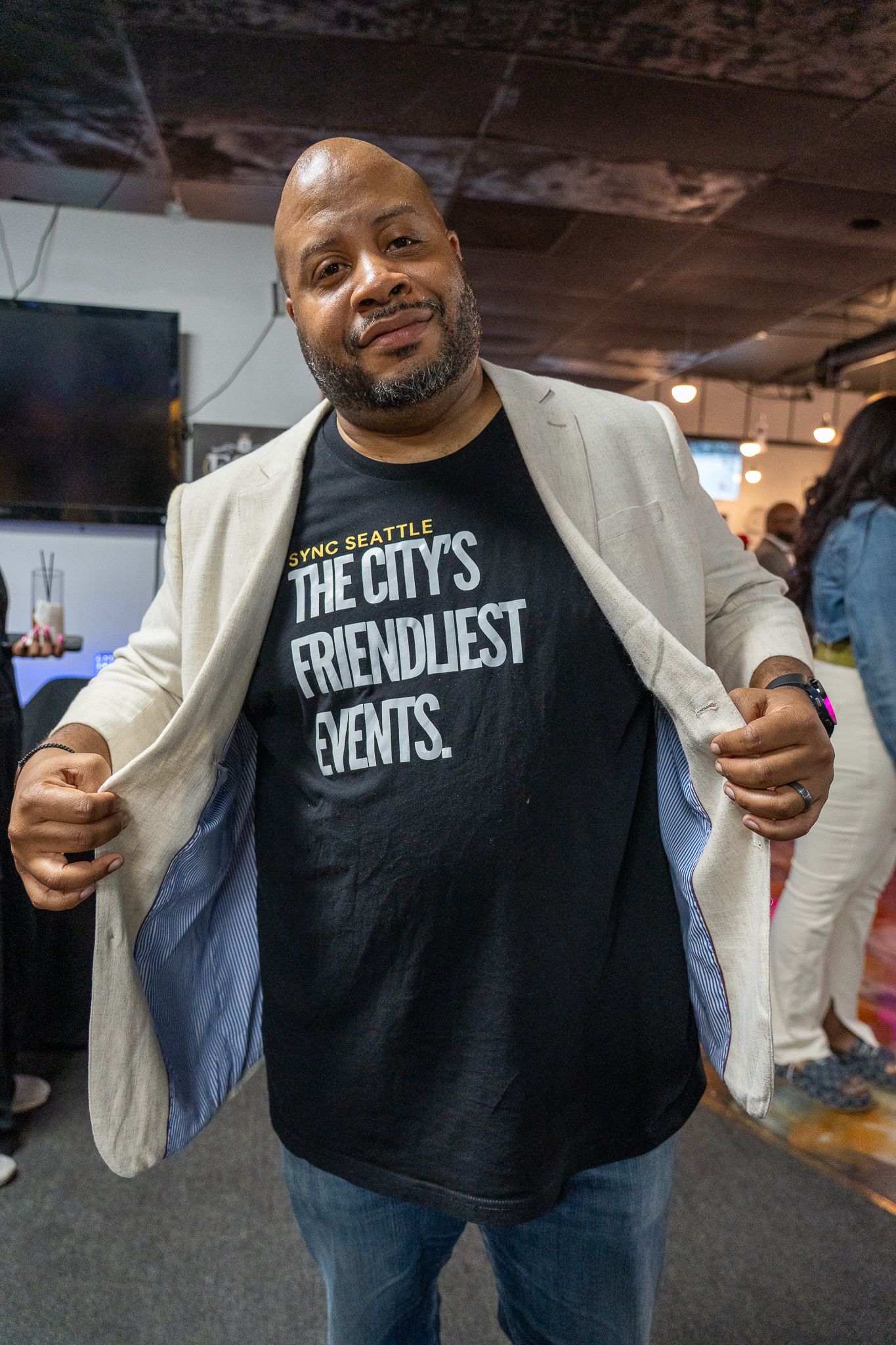 A person wearing a black t-shirt with 'The City's Friendliest Events' text and light blazer gestures enthusiastically.