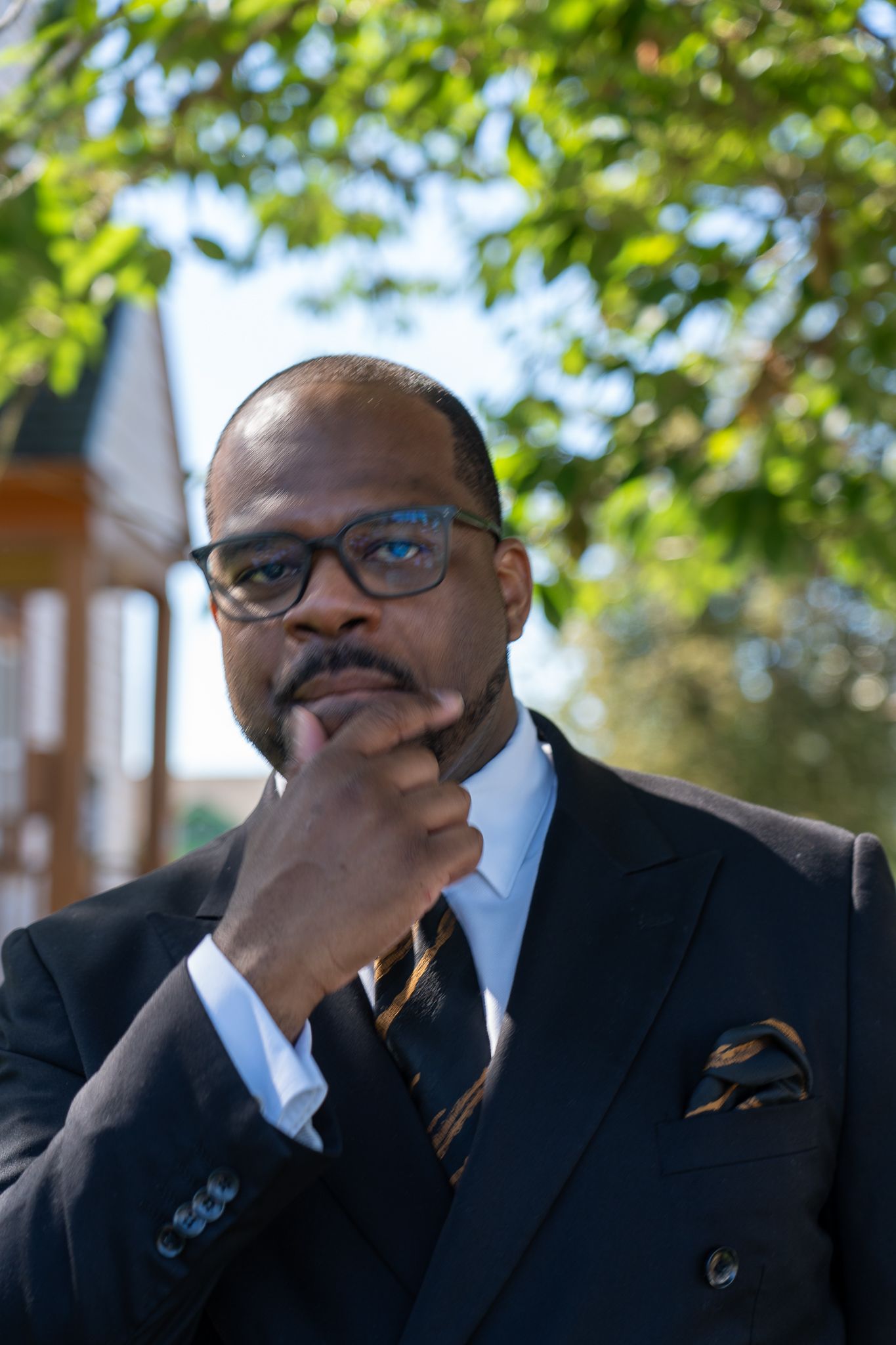 Professional portrait of someone in a dark business suit with glasses and pocket square in thoughtful pose outdoors.