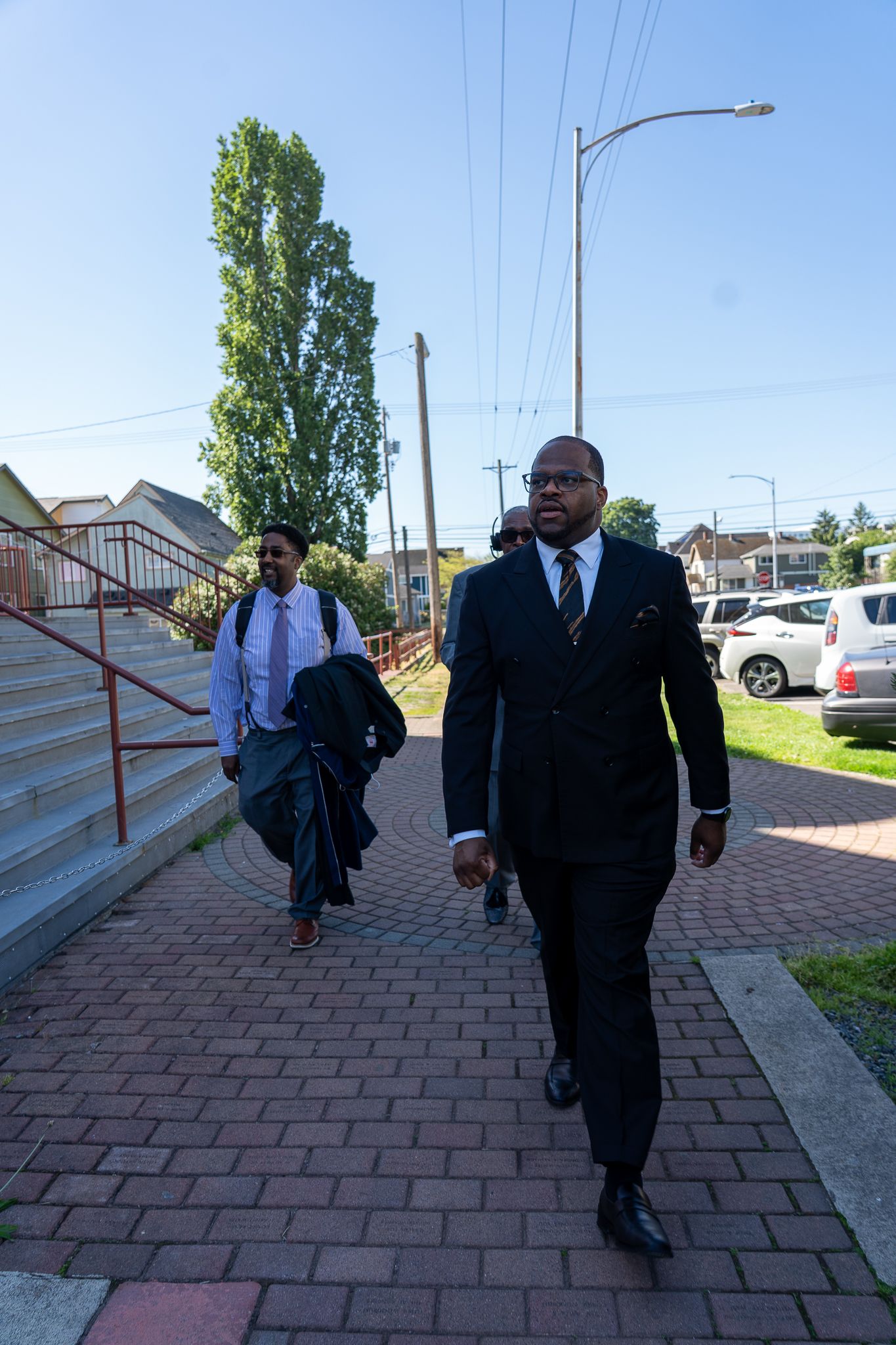 People in formal business attire walking along a brick paved sidewalk on a sunny day with trees and buildings in background.