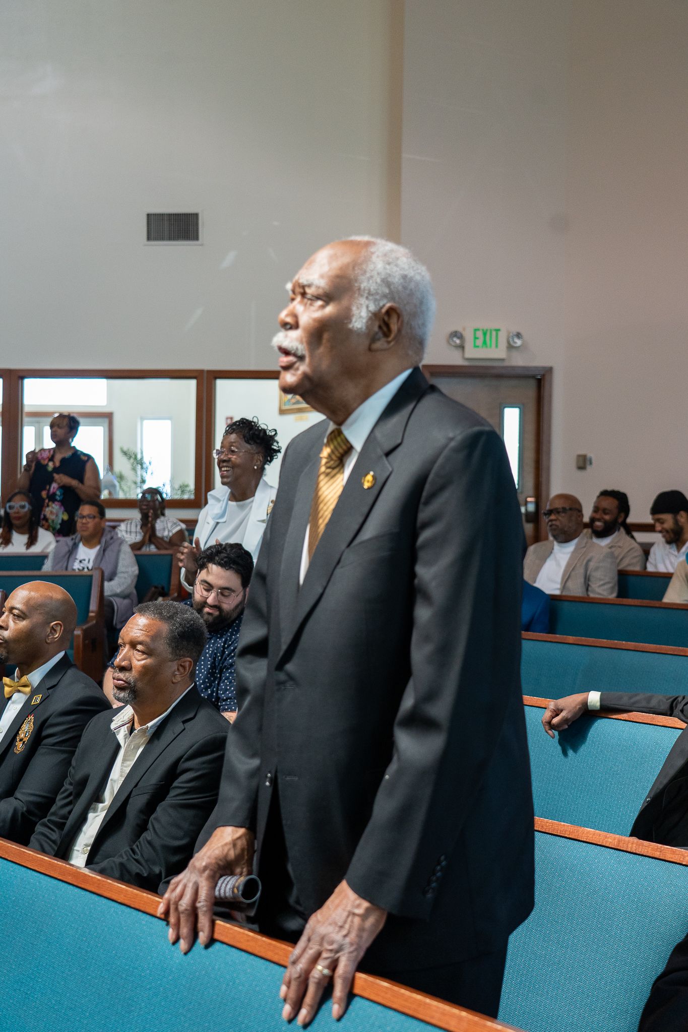 Inside a church sanctuary, members of the congregation stand during a service in turquoise pews.