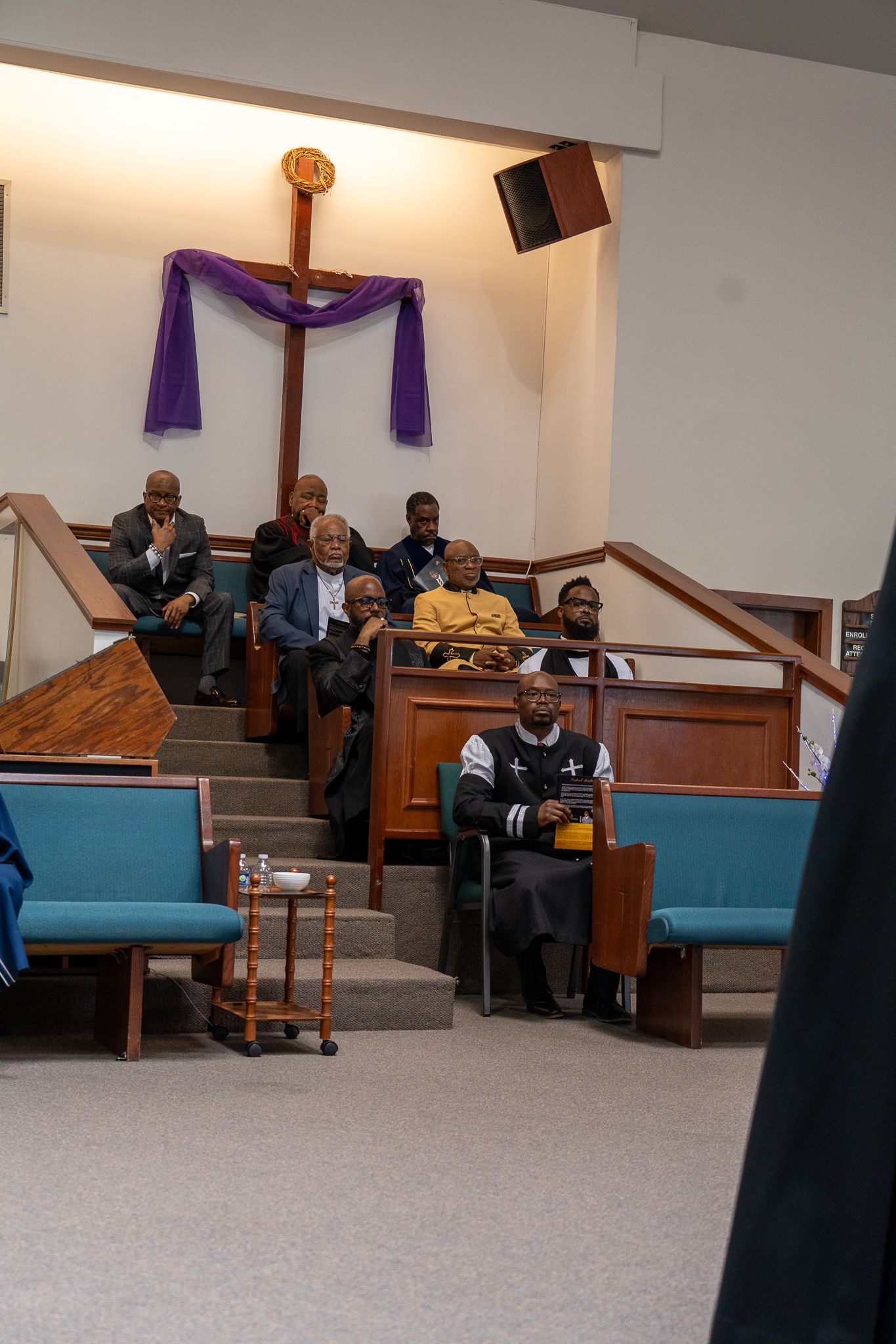 Several people sit in pews inside a church or chapel with teal-colored bench cushions and purple decorative elements.