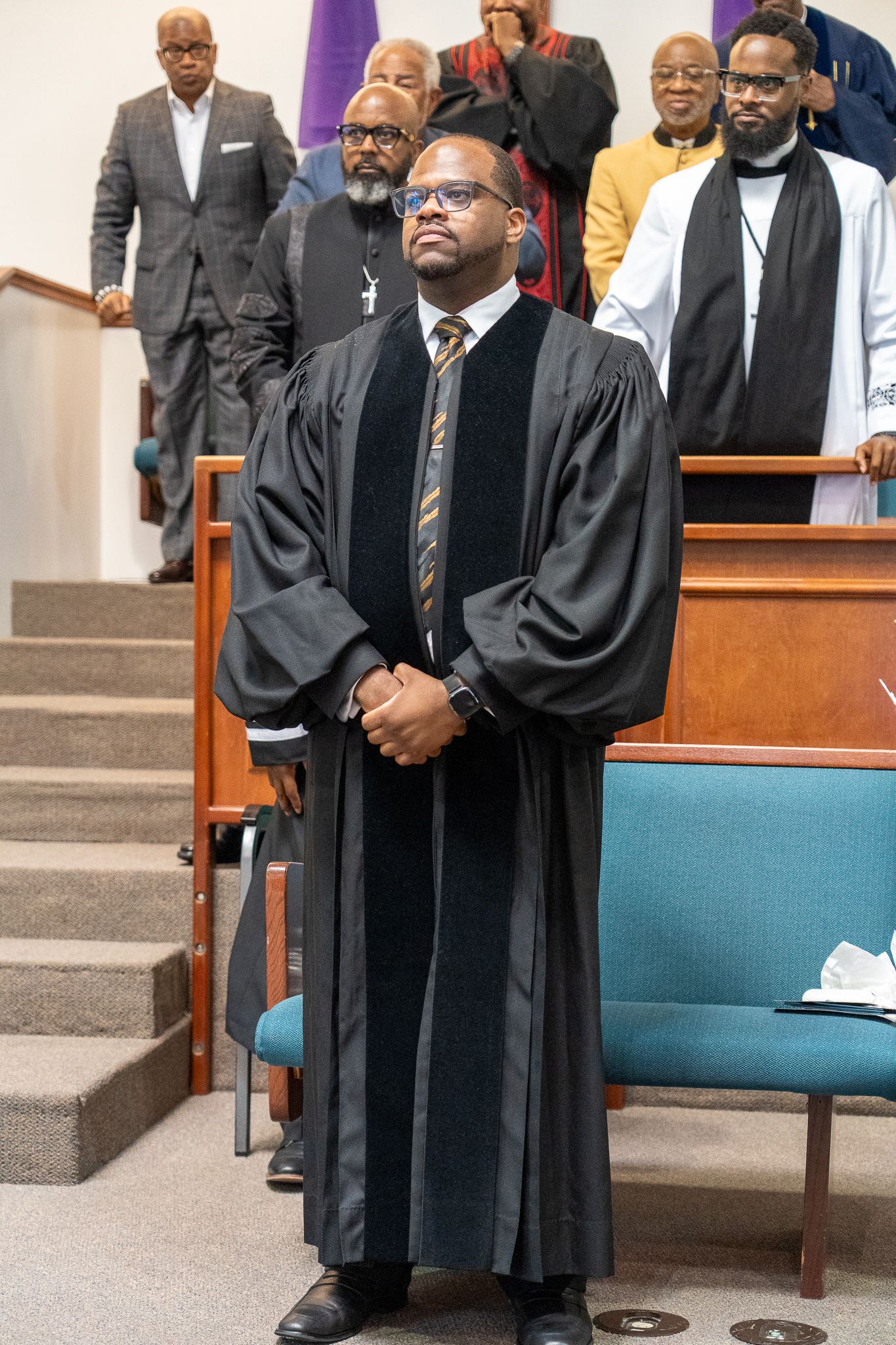 Several people in black graduation gowns and formal attire stand and wait in a church or auditorium setting.
