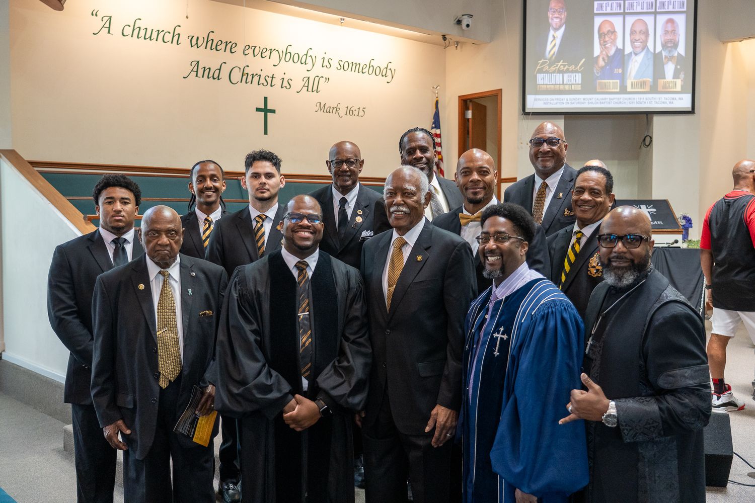 A group of formally dressed individuals in suits and graduation gowns gather beneath an inspirational quote on a wall.