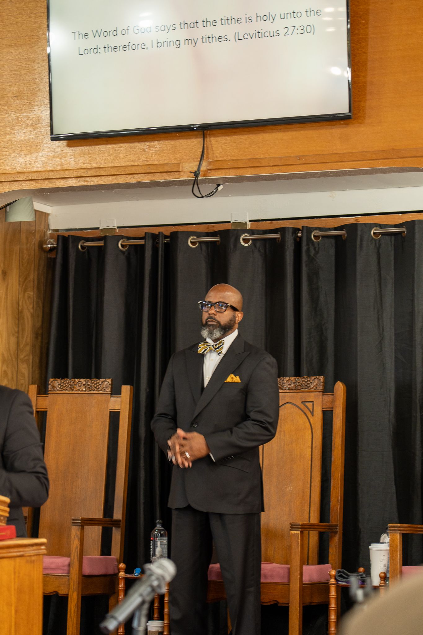 A speaker stands at a church podium wearing a black suit with yellow pocket square, with scripture text visible above.
