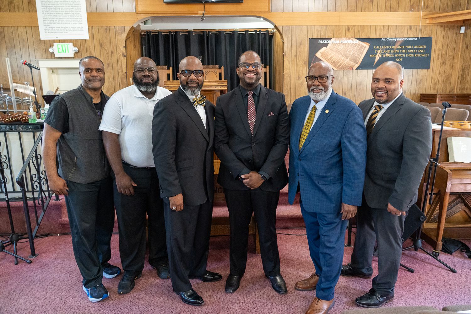 A group of six men wearing suits and formal attire stand together smiling in what appears to be a church or religious setting.